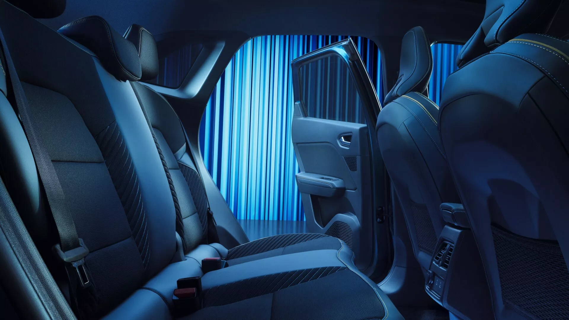 Obrázek zobrazuje interiér vozu Renault Symbioz 2024, s důrazem na zadní sedadla z úhlu uvnitř kabiny vozu, mírně směrem k straně spolujezdce. Pohled je nízký, v blízkosti úrovně sedadla, detailně zobrazující materiál sedadel, prošívání a design s modrým ambientním osvětlením zvýrazňujícím kontury. Viditelné jsou rozvržení zadních sedadel s opěrkami hlavy a bezpečnostními pásy, část předního sedadla spolujezdce, potažená kvalitní látkou nebo kůží pro luxus a pohodlí. Dveřní panel je osvětlen modrým světlem a obsahuje ovládací tlačítko, možná na okna nebo jiné pokročilé funkce, spolu s mřížkou reproduktoru a úložným prostorem. Tmavé koberce svědčí o praktičnosti. Obrázek naznačuje funkce jako pokročilé ambientní osvětlení, prémiové materiály, elektronické ovládání a možná prémiový zvukový systém, což odráží klidný, moderní interiér zaměřený na pohodlí pasažéra.
