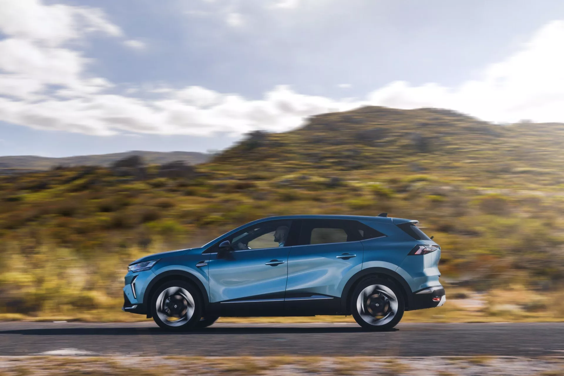 Jasně modrý Renault Symbioz 2024 (1. generace) v pohybu, ukazující své elegantní aerodynamické linie a moderní design z profilu. Auto je umístěno v levé třetině obrázku, má rozmazané pozadí a silnici, což naznačuje rychlost. Viditelné prvky zahrnují LED denní svícení, integrované směrovky ve zpětných zrcátkách, panoramatickou střechu, výrazné ráfky a nízkoprofilové pneumatiky. Fotografie je pořízena technikou panning, která zdůrazňuje dynamický pohyb auta.