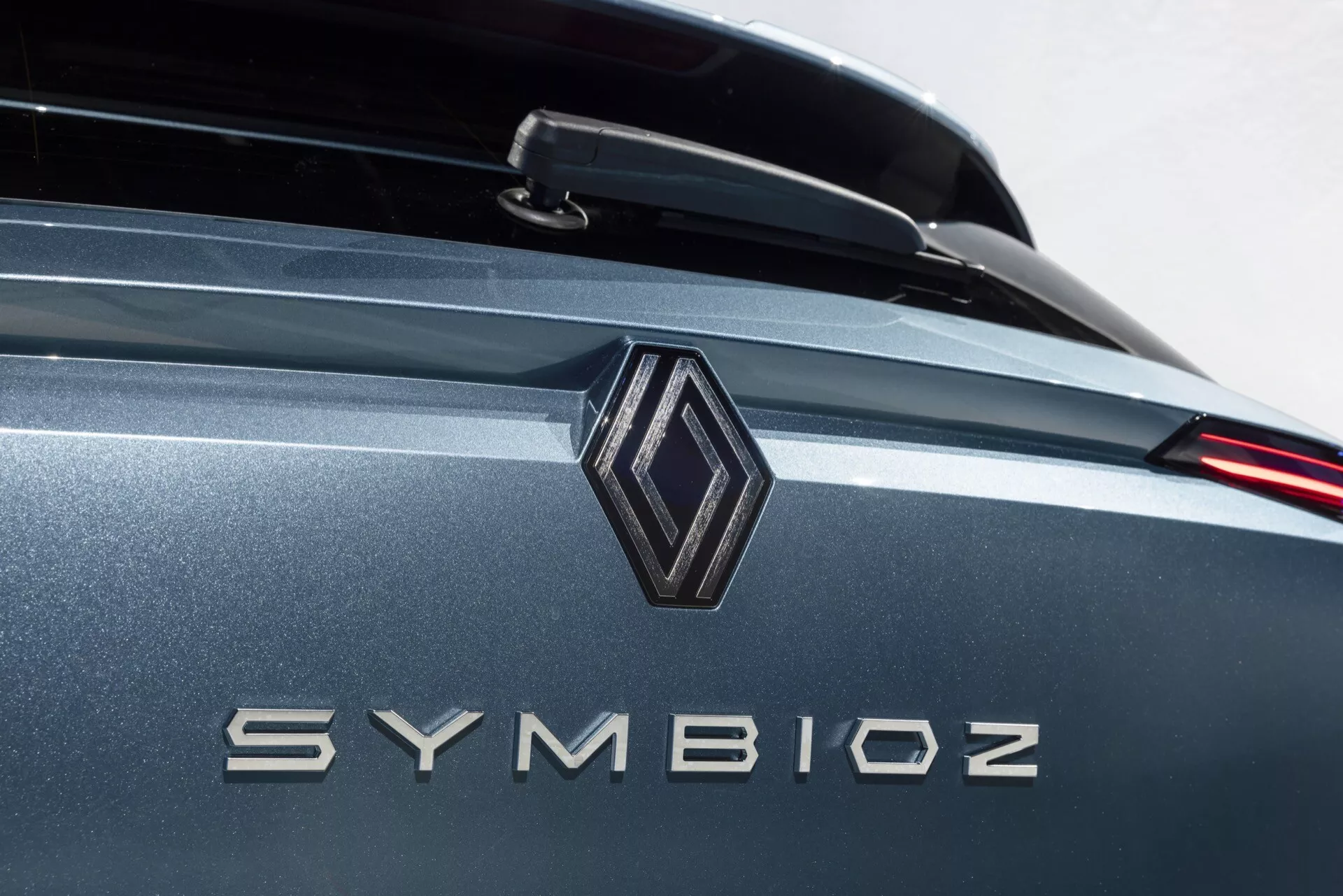 Profesionální detailní snímek zadní části auta zaměřující se na brandingové detaily s diamantovým emblémem a názvem modelu 'SYMBIOZ' v kovových písmenkách. Obrázek využívá malou hloubku ostrosti, což zvýrazňuje emblém a nápis, zatímco pozadí jemně mizí. Dobře osvětlené prostředí naznačuje vnitřní showroom nebo studio. Barva vozidla je kovově šedá nebo modrá s reflexními vlastnostmi, obsahující možný zadní spoiler a moderní design LED koncových světel.