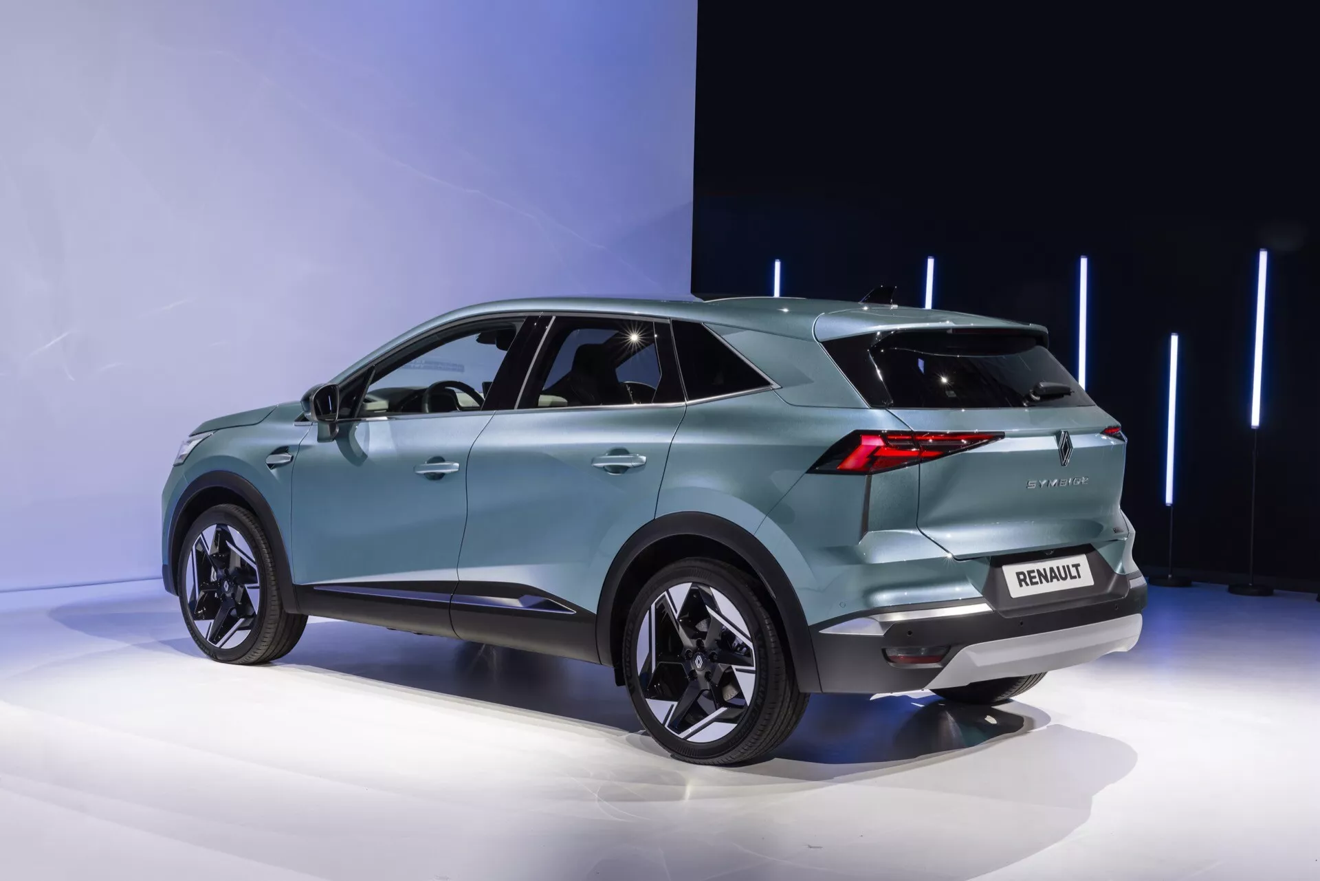 Tříčtvrteční zadní pohled na Renault Symbioz 2024. Tyrkysové SUV, umístěné na výstavní plošině, ukazuje moderní aerodynamický design se svažující se střechou, výraznými blatníky, které obsahují velké hliníkové kola, a tónovanými zadními okny. Výrazné LED zadní světla, zadní spoiler, logo na zadní straně a označení modelu, parkovací senzory a pravděpodobně zadní kameru jsou viditelné. Elegantní zadní nárazník také obsahuje světelné prvky, zatímco žraločí ploutev naznačuje pokročilou technologii. Integrované ukazatele směru ve vnějších zrcátkách a zapuštěné kliky dveří naznačují bezklíčový vstup. Minimalistické pozadí zvýrazňuje detaily vozu.
