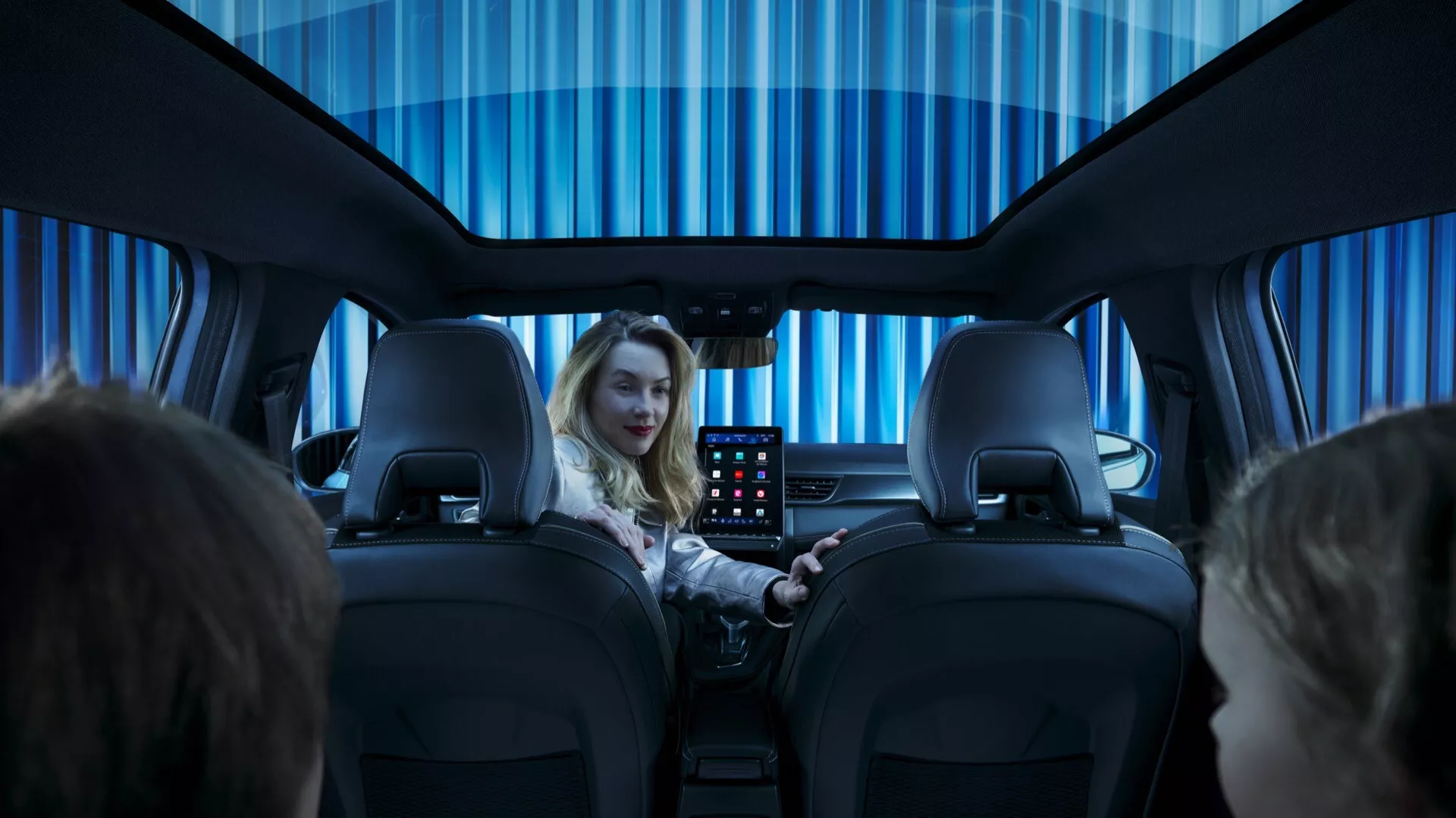 Interiér Renault Symbioz 2024, první generace, fotografovaný ze zadního sedadla směrem dopředu. Ukazuje zadní část dvou předních sedadel s nastavitelnými opěrkami hlavy, ženu na sedadle řidiče, která se směje směrem ke kameře, a interaktivní displej umístěný mezi sedadly pro zadní cestující. Rovnoměrně osvětlený, studeně modrý interiér naznačuje moderní, luxusní atmosféru, zvýrazněnou tónovanou panoramatickou střechou.