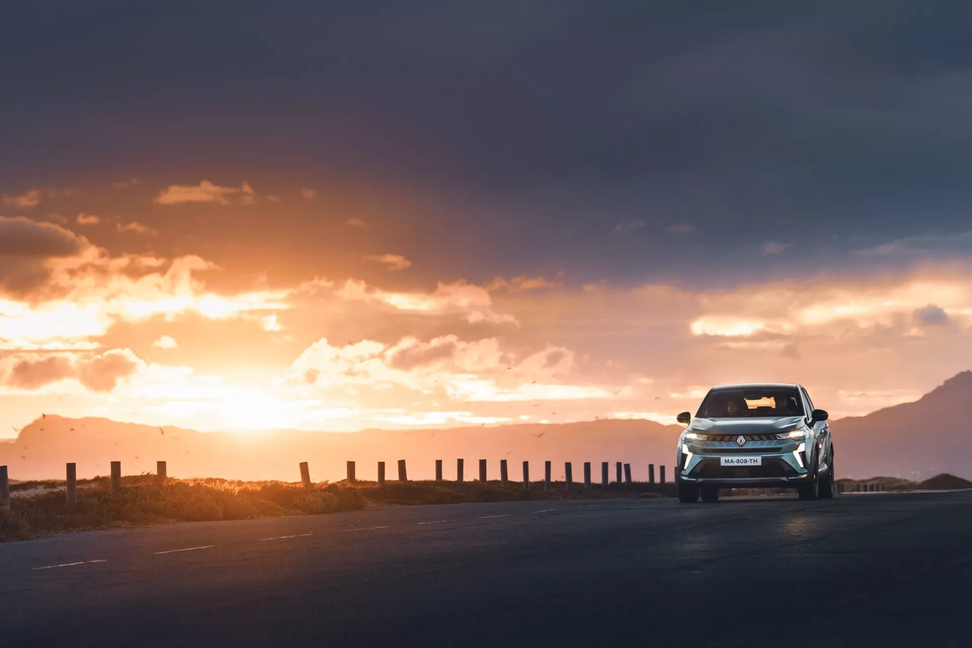 Obrázek zachycuje Renault Symbioz 2024 z nízkého úhlu, což zdůrazňuje jeho dynamickou a silnou přítomnost při jízdě směrem k fotoaparátu. Fotografie ukazuje celý předek vozu, s viditelnou částí boků, osvětlený teplým, zlatavým protisvětlem zapadajícího slunce. Světlomety a denní světla jsou zapnuté, což zvýrazňuje agresivní design zamýšlený pro tento novější model. Navzdory siluetovému efektu zůstávají viditelné nárazník, mřížka chladiče, kapota a čelní sklo. Vozidlo se nachází na prázdné zpevněné silnici ohraničené plotovými sloupky, pod dramatickou oblohou s rozptýlenými mraky, což zvyšuje pocit dobrodružství a objevování.