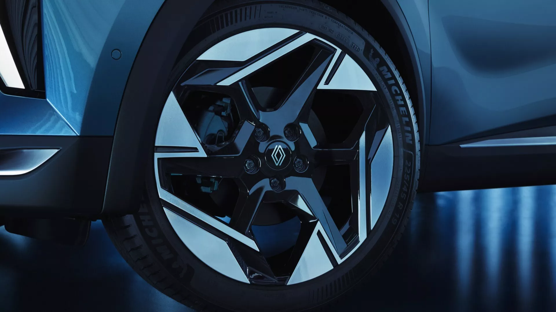 Detailní pohled na přední kolo a částečný boční profil Renault Symbioz 2024 (1. generace) pořízený z nízkého úhlu. Slitinové kolo má složitý, geometrický vícepaprskový design s lesklým a matným provedením, spárováno s nízkoprofilovými výkonnostními pneumatikami a značkovými třmeny, což naznačuje sportovnost a luxus. Studený modrý tón zvyšuje estetiku, naznačuje studiové osvětlení nebo postprocessing.