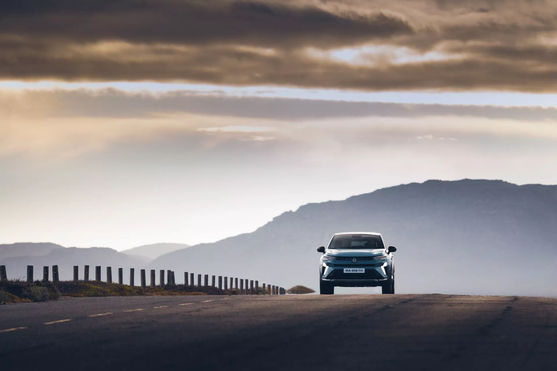 Přední úhel pohledu na vůz Renault Symbioz 2024 (1. generace), zachycený během brzkého rána nebo pozdního odpoledne s teplým osvětlením a dlouhými stíny. Vozidlo s elegantními liniemi designu a výraznými světlomety se pohybuje směrem ke kameře na vysoko položené silnici, pozadí s horami mírně rozostřené. Osvětlená oblačná obloha dodává atmosférický kontext; nízká perspektiva zdůrazňuje přední části jako mřížku, kapotu a nárazník. Pohybové rozmazání na okrajích ukazuje auto v pohybu při zachování ostrého zaostření na moderní techniku a design.