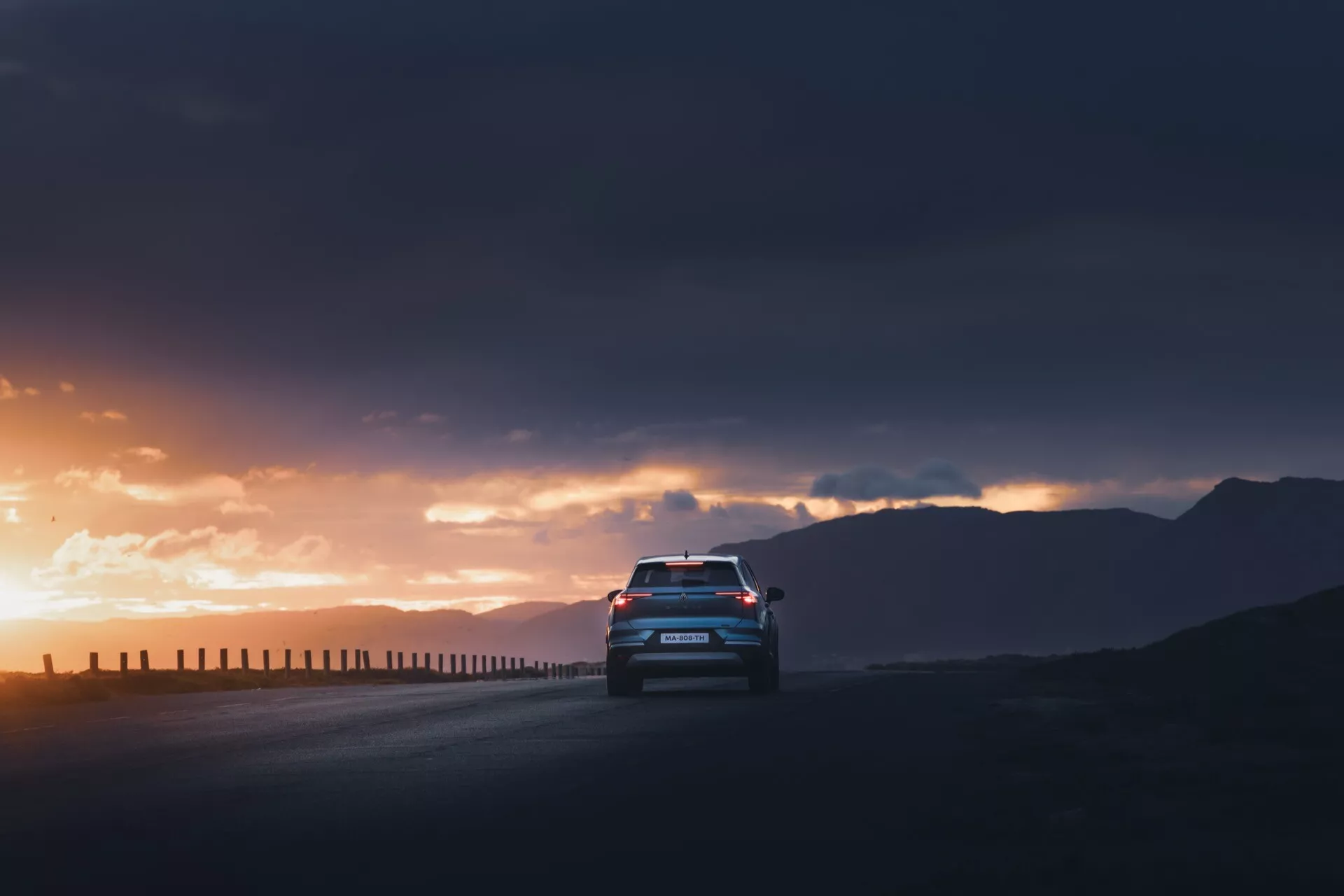 Zadní pohled z nízkého úhlu na Renault Symbioz 2024, 1. generace, jedoucí po silnici proti dramatickému východu nebo západu slunce. Silueta vozu je zvýrazněna osvětlenými LED zadními světly a zadními designovými prvky jako spoiler, dvojitý výfukový systém a zadní difuzor. Tmavá, zamračená obloha s paprsky slunce propouštějícími skrze mraky zvýrazňuje přítomnost vozidla, zatímco odrazy na zadním okně naznačují interiérové osvětlení nebo ambientní světlo.