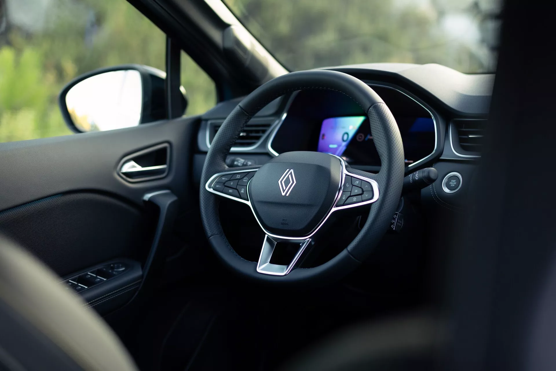Pohled do interiéru vozu Renault Symbioz 2024 z místa na zadním sedadle, zaměřený na moderní třípaprskový volant a digitální přístrojovou desku. Volant má ploché dno, kovový akcent a integrovaná ovládací tlačítka, vše v temném barevném schématu. Mělká hloubka ostrosti zdůrazňuje tyto prvky, rozmazané okolí včetně opěrky hlavy a venkovní scenérie viditelné skrz okno naznačuje, že vůz je v klidovém stavu.