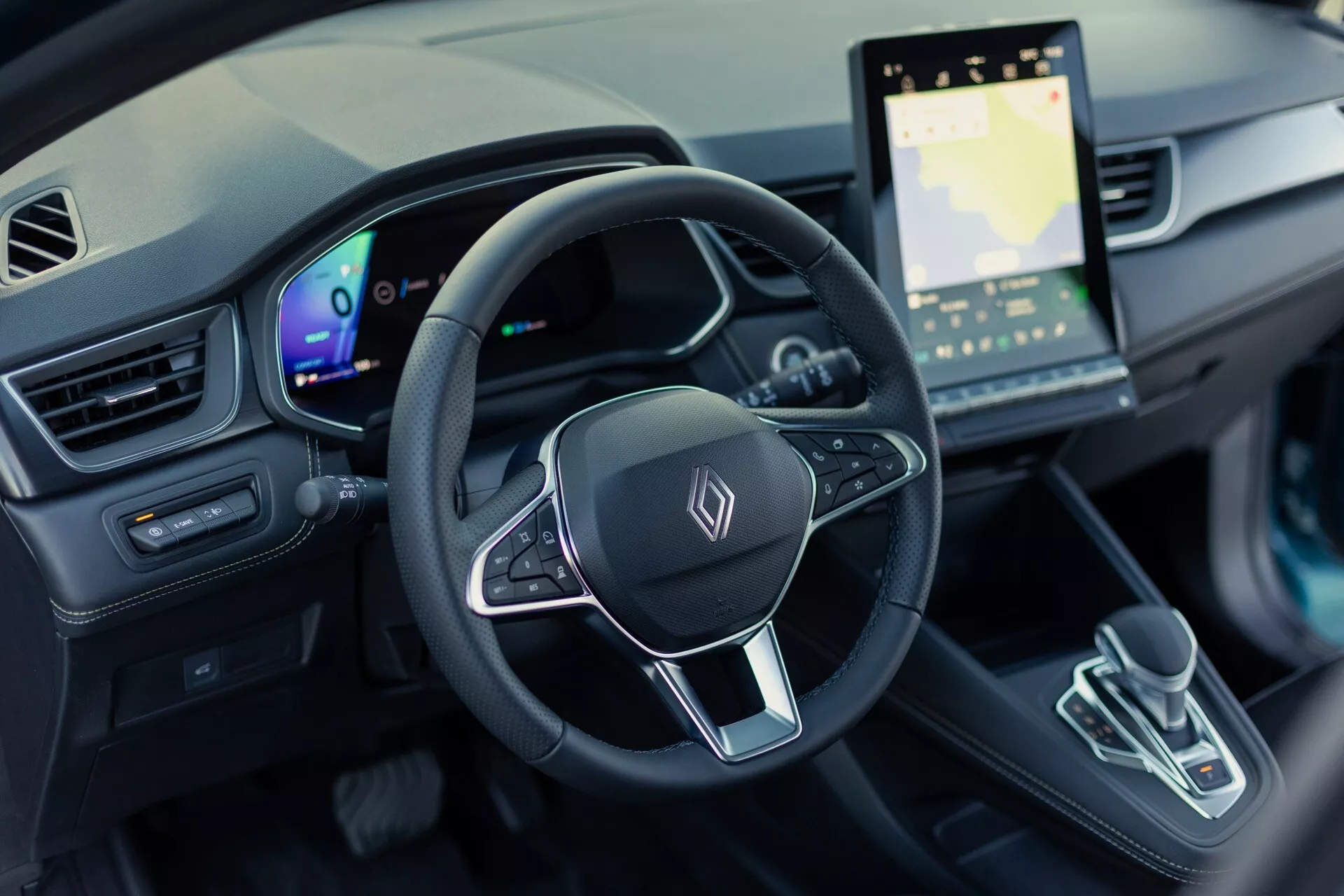 Záběr interiéru Renault Symbioz 2024 (1. generace) z pohledu řidiče, zaměřený na volant a palubní desku. Obrázek poskytuje pohled z ptačí perspektivy, 30 stupňů dolů, a zobrazuje tříramenný volant s multifunkčními tlačítky a obecným emblémem, plně digitální přístrojový štít a velký dotykový infotainmentový systém se zobrazením mapy. Středová konzola zahrnuje moderní volič převodovky s minimalistickým designem. Interiér představuje vysoce technologický design elektrického vozidla s ambientním osvětlením a moderní estetikou.