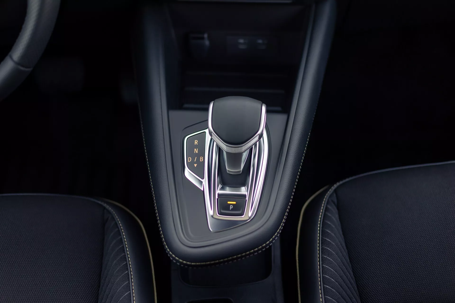 Pohled shora na středovou konzolu Renault Symbioz 2024 se zaměřením na elektronický volič převodovky s chromovaným povrchem a osvětlenými indikátory převodů pro parkování, zpátečku, neutral a jízdu. Volič má kožené boky, žlutě zvýrazněné tlačítko parkování a je umístěn v lesklém černém pouzdře s propracovaným prošitím okolního koženého interiéru.