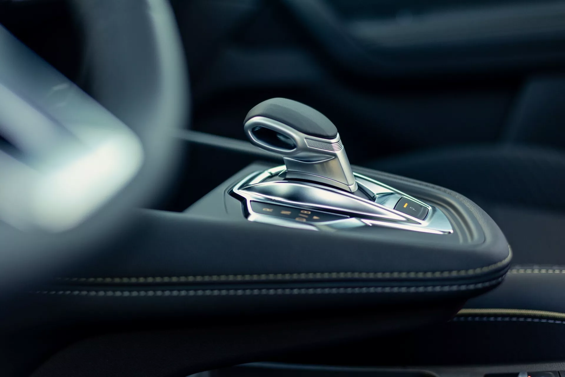 Detailní interiérový záběr řadicí páky Renault Symbioz 2024, který ukazuje moderní design s kovovými akcenty a pravděpodobně koženým povrchem. Vysoce rozlišený snímek pořízený z nízkého úhlu se zaměřuje na automatickou řadicí páku a okolní středovou konzoli, která je vyrobena z vysoce kvalitních materiálů, jako je kůže, broušený kov nebo chrom. Na konzoli jsou viditelné různé tlačítka pravděpodobně pro jízdní režimy, parkovací brzdu, funkci auto-hold a kontrolu trakce. Měkké, přirozené osvětlení zdůrazňuje luxusní a precizní inženýrství interiéru vozu.