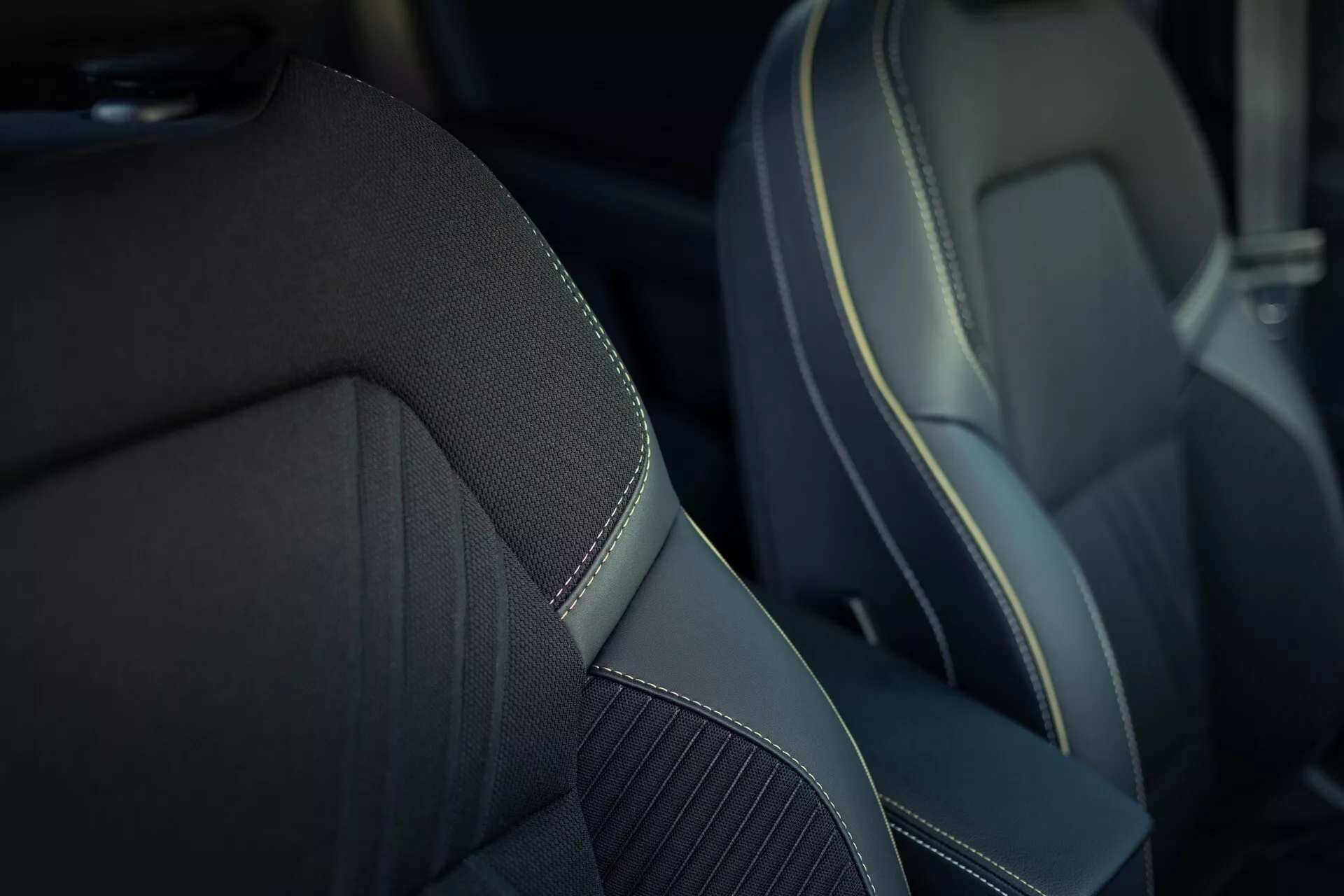 Detailní pohled na přední sedadlo spolujezdce v Renaultu Symbioz 2024 (1. generace). Sedadlo má střední panel vyrobený z texturované látky nebo syntetického tkaniva pro lepší prodyšnost, obklopený hladkým syntetickým materiálem podobným kůži s kontrastním ozdobným šitím, které zvýrazňuje prémiový nebo sportovní design. Mělká hloubka ostrosti se zaměřuje na sedadlo a rozmazává pozadí, aby vynikla kvalita materiálu a řemeslné zpracování.