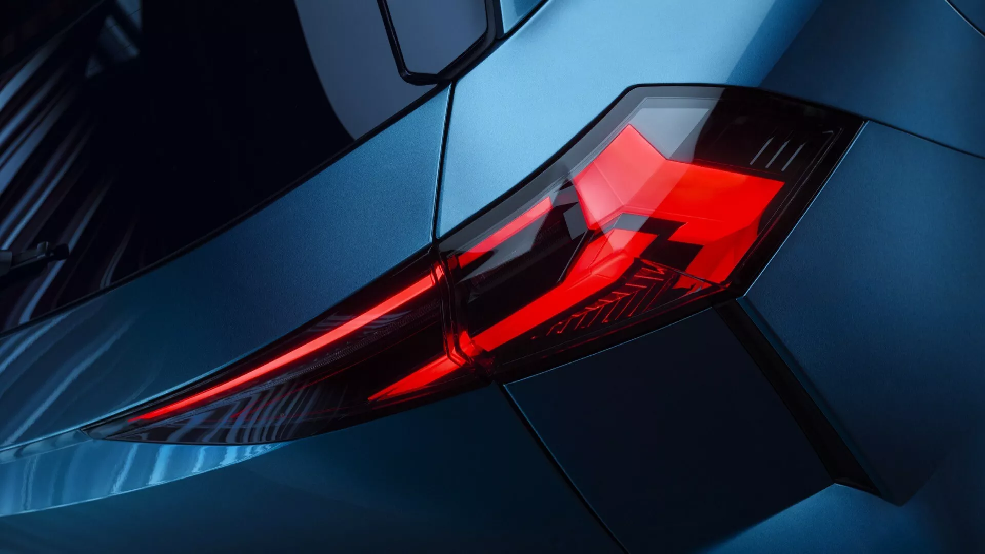 Detailní pohled na zadní světlo automobilu Renault Symbioz 2024, zdůrazňující LED osvětlovací prvky v úhlovém, vrstveném designu krytu. Fotografie je pořízena z šikmého úhlu, zaměřuje se na zadní levý roh s vyvýšeným pohledem, zvýrazňující kovově modrou karoserii vozu a aerodynamické linie. Obrázek zdůrazňuje moderní, sportovní design a prémiovou estetiku vozidla, vhodné pro diskuse na téma automobilové technologie a designové trendy.