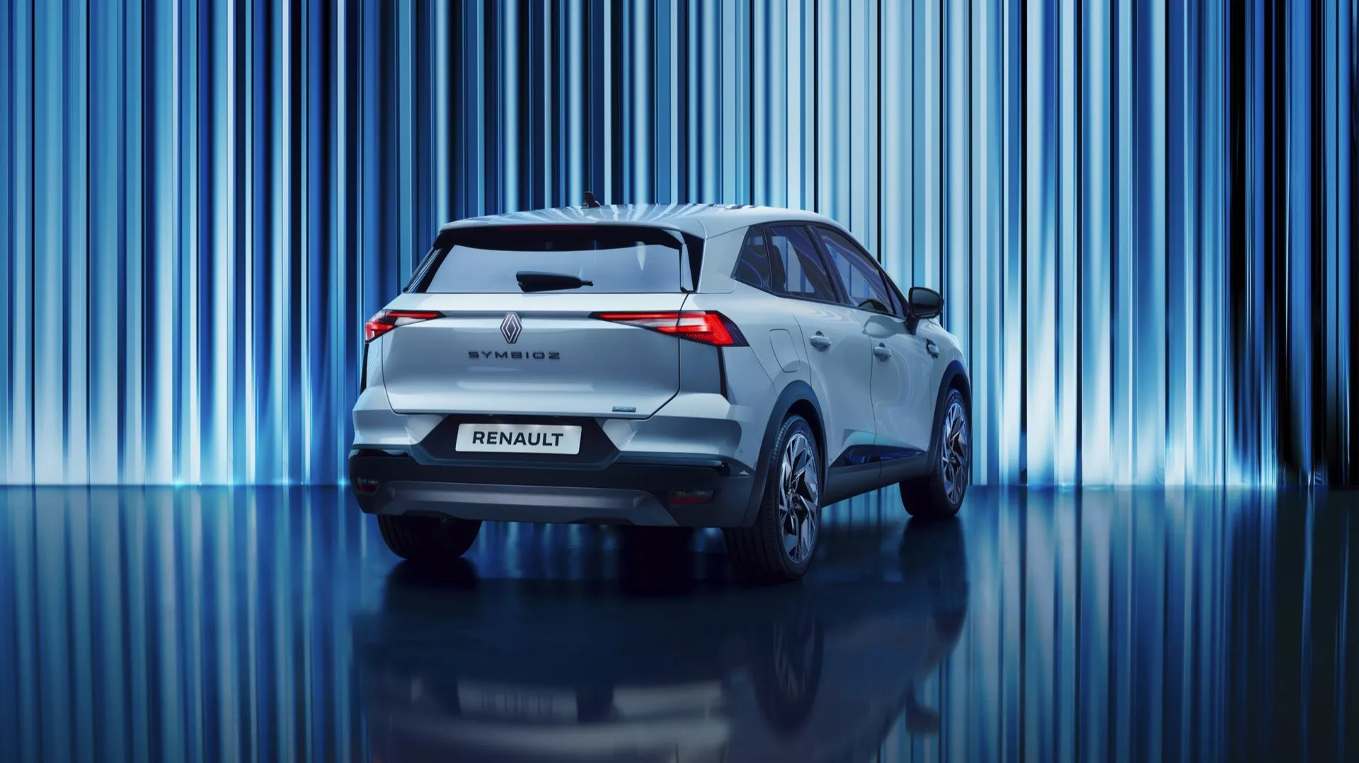 Tento profesionální snímek ukazuje zadní část prvního modelu Renault Symbioz 2024, moderní SUV. Vozidlo má charakteristická LED zadní světla, zadní spoiler, zadní stěrač, znak Renault a název modelu. Záběr z nízkého úhlu zdůrazňuje jeho postoj a zvýrazňuje čistý design bez viditelného výfuku, což naznačuje hybridní nebo elektrický pohon. Na živém pozadí s vertikálními modrými světelnými pruhy, lehké metalické odstíny vozu a reflexní podlaha vytvářejí sofistikovanou atmosféru showroomu či inscenované fotoakce.