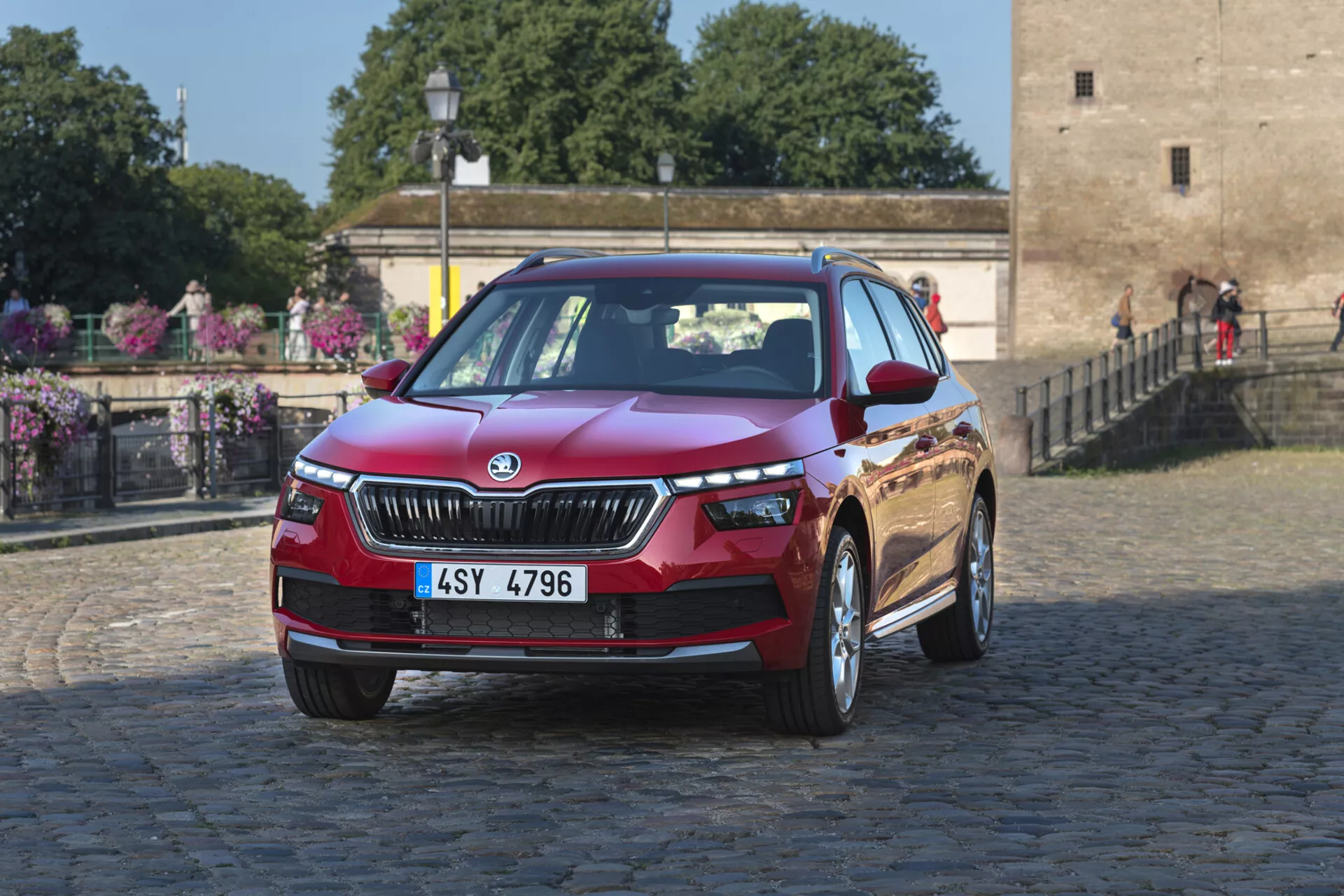 Červený Škoda Kamiq z roku 2019, fotografován z předního tříčtvrtečního úhlu, ukazuje přední mřížku, LED světlomety, kapotu, přední nárazník s možnými mlhovými světly nebo vzduchovými vstupy a slitinová kola, na pozadí městského nebo historického bruku s vegetací.