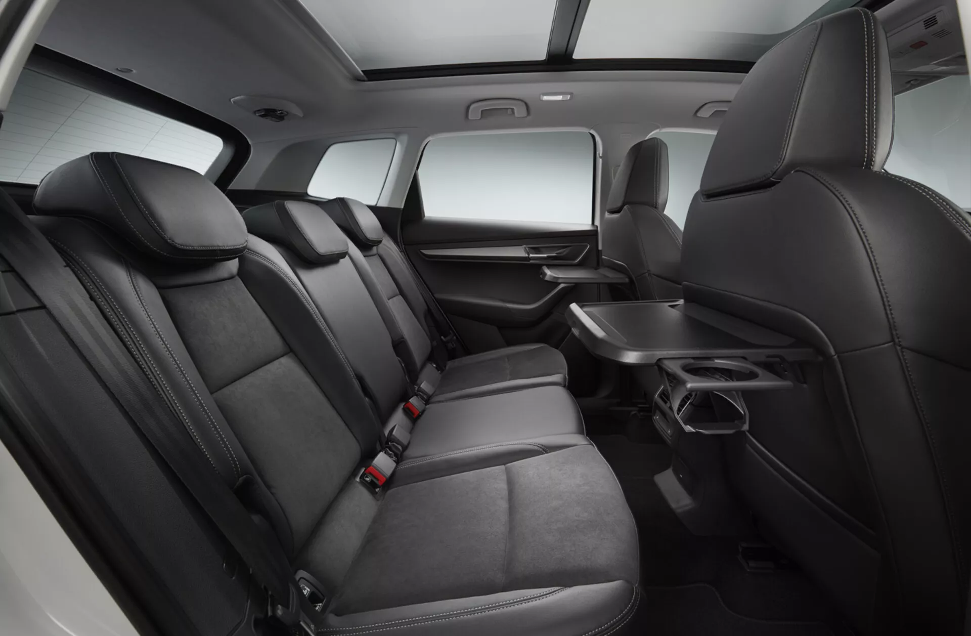 Interiérový pohled na Skoda Karoq 2017 zaměřený na prostor druhé řady sedadel s prémiovými černými koženými sedadly, sklopným opěradlem s držáky na nápoje, dveřním panelem s ovládáním elektriky, kapsami za předními sedadly, bezpečnostními pásy pro všechny pozice a konzolí uprostřed rozšiřující se do druhé řady, vše pod zvýrazněným studijským osvětlením