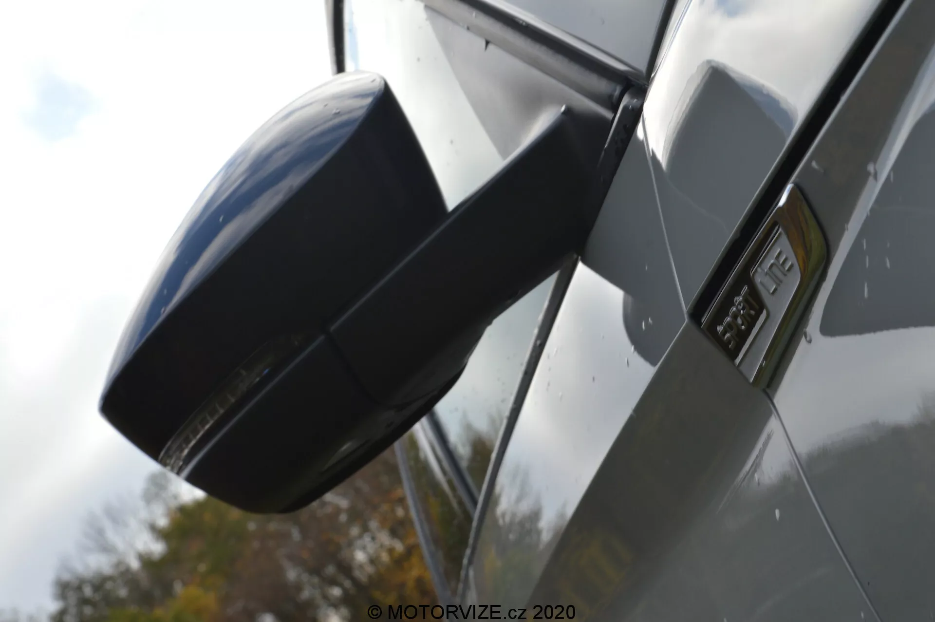 Detailní pohled z boku na Škoda Kodiaq 2017 se zaměřením na oblast zrcátka a blatníku. Obrázek je pořízen z nízkého úhlu, čímž zdůrazňuje boční profil vozu, s moderním aerodynamickým zrcátkem barvou sladěným s karoserií, výrazným znakem na blatníku indikujícím konkrétní model nebo výbavový stupeň, lesklými kovovými panely karoserie a odrazy, které naznačují venkovní prostředí s tmavým vozem.