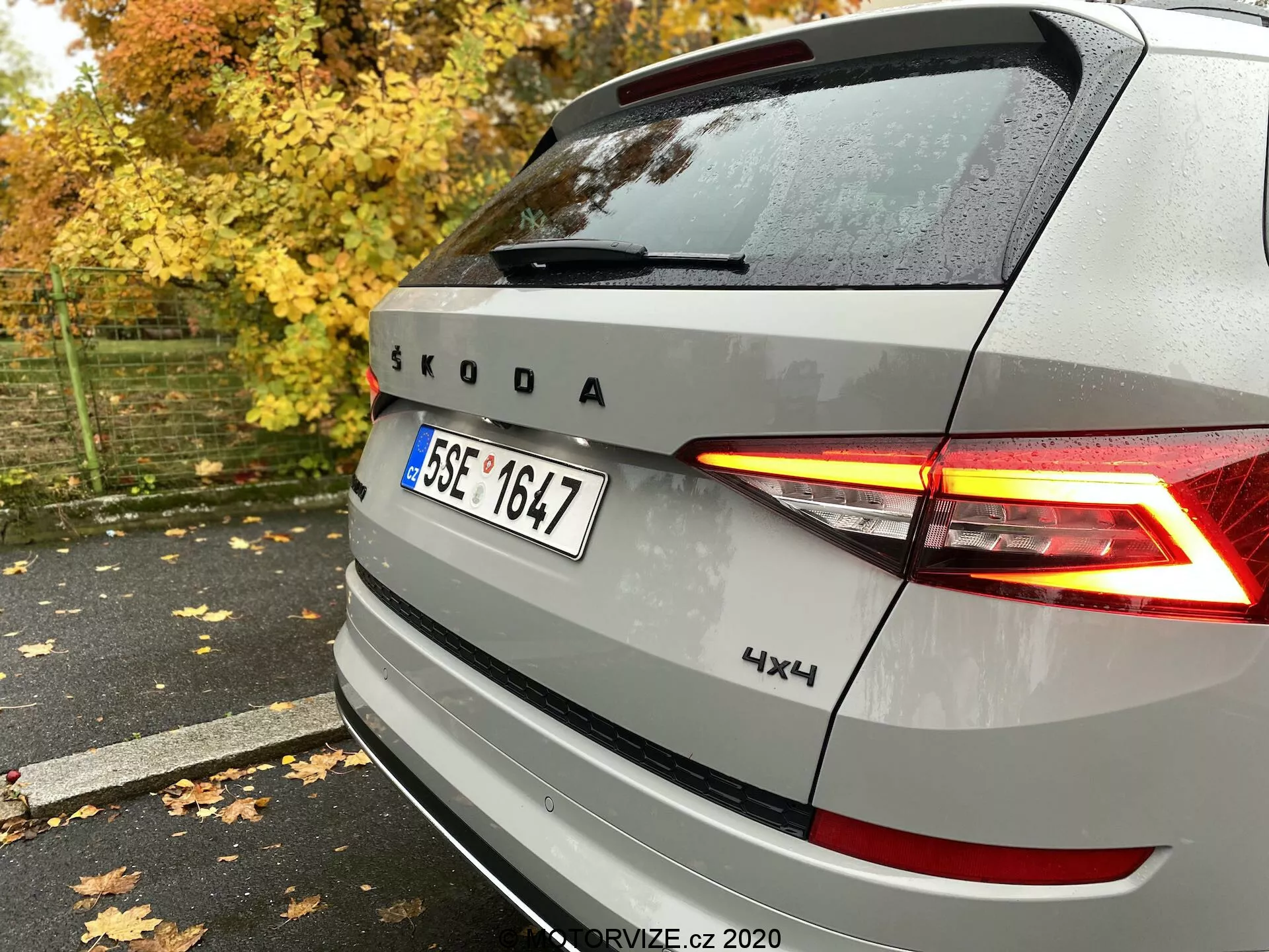 Zadní pohled na stříbrné vozidlo Škoda s osvětlenými LED zadními světly ve tvaru C, znakem Škoda a značkou, s označením 4x4, zachycený zezadu zprava na mokrém chodníku mezi podzimními listy a stromy.