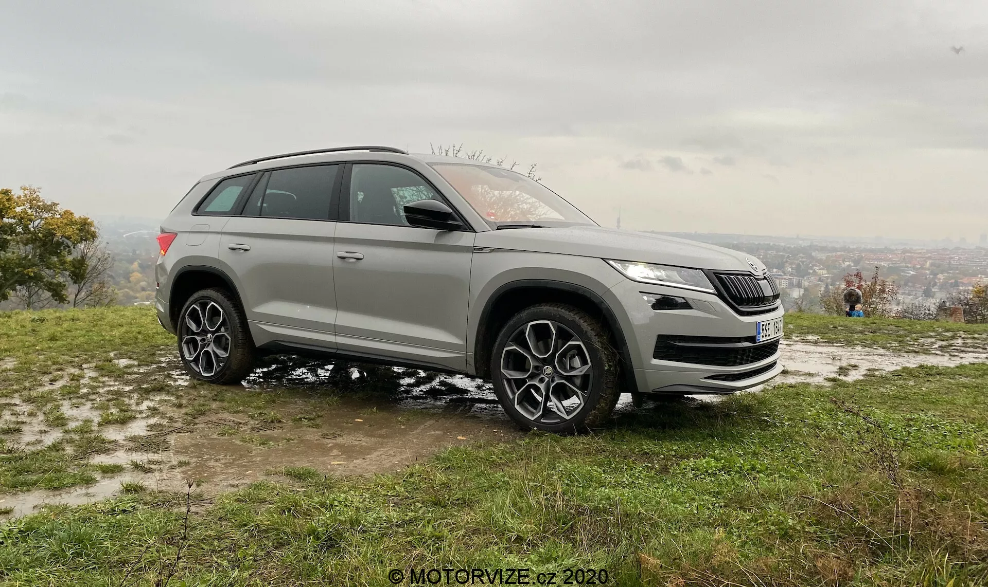Stříbrné SUV Skoda Kodiaq 2017 zaparkované na trávnatém a mírně blátivém povrchu, zaznamenané zbočního předního úhlu. Jasný pohled na přední masku, světlomety, levý boční profil, elegantní design světlometů s LED technologií, výrazný design masky, senzory pro asistenční systémy mohou být viditelné pod maskou, boční zrcátka s ukazateli směru a možnou detekcí mrtvého úhlu, velká kola z vícepaprskové slitiny, pneumatiky vhodné pro silniční i mírné off-roadové podmínky, naznačující možnosti pohonu všech kol. Umístěno na svahu s oparovým pohledem na město v pozadí pod zataženou oblohou, měkké osvětlení naznačující časné ráno nebo pozdní odpoledne. Lidé v pozadí užívající výhled, zvýrazňující rekreační a životní stylový nádech.
