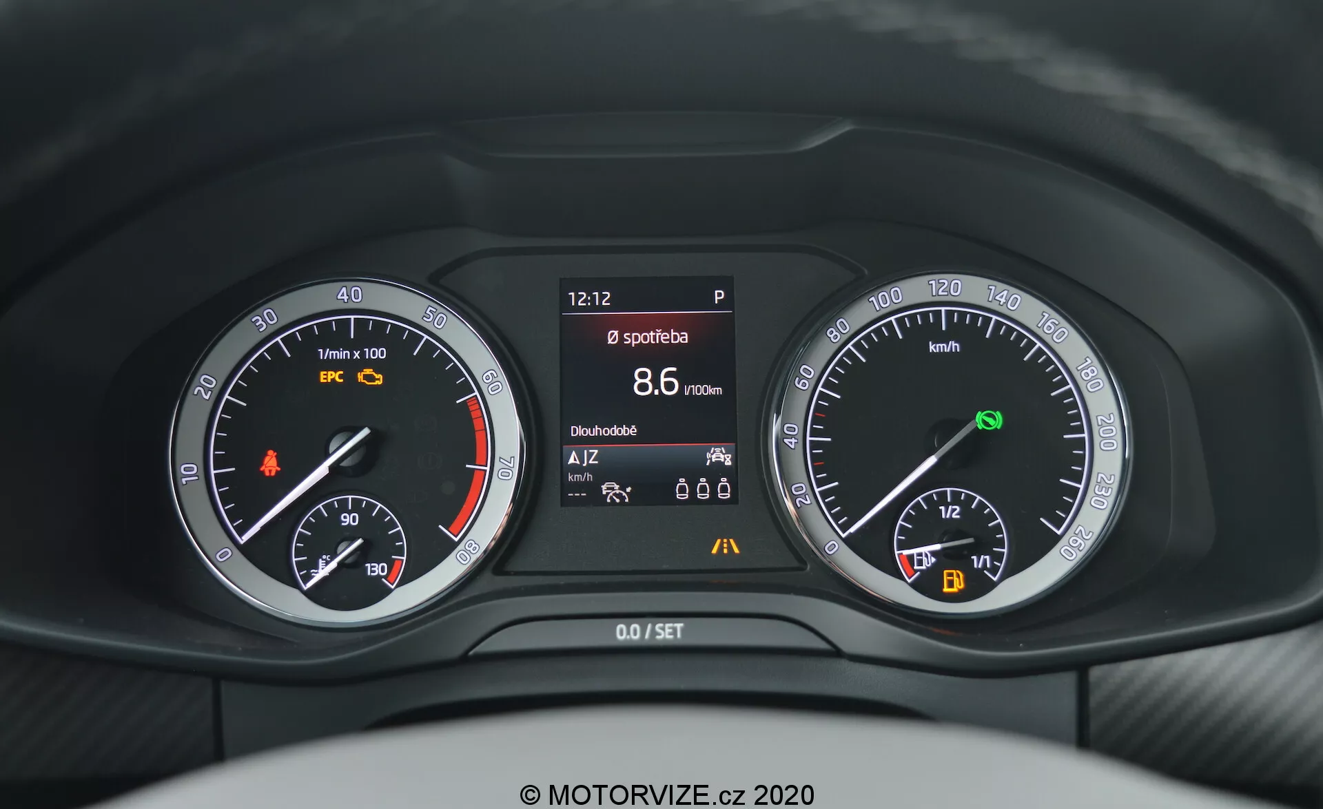 Detailní záběr z pohledu řidiče na přístrojovou desku vozu Škoda Kodiaq 2017, která kombinuje analogové a digitální ukazatele za volantem. Přístrojová deska obsahuje otáčkoměr s oranžovou kontrolkou, centrální obrazovku zobrazující různé informace o vozidle, jako je čas, stav parkovací brzdy, sportovní režim, dojezd a teplota, a rychloměr s ukazatelem paliva a indikátorem start/stop systému.