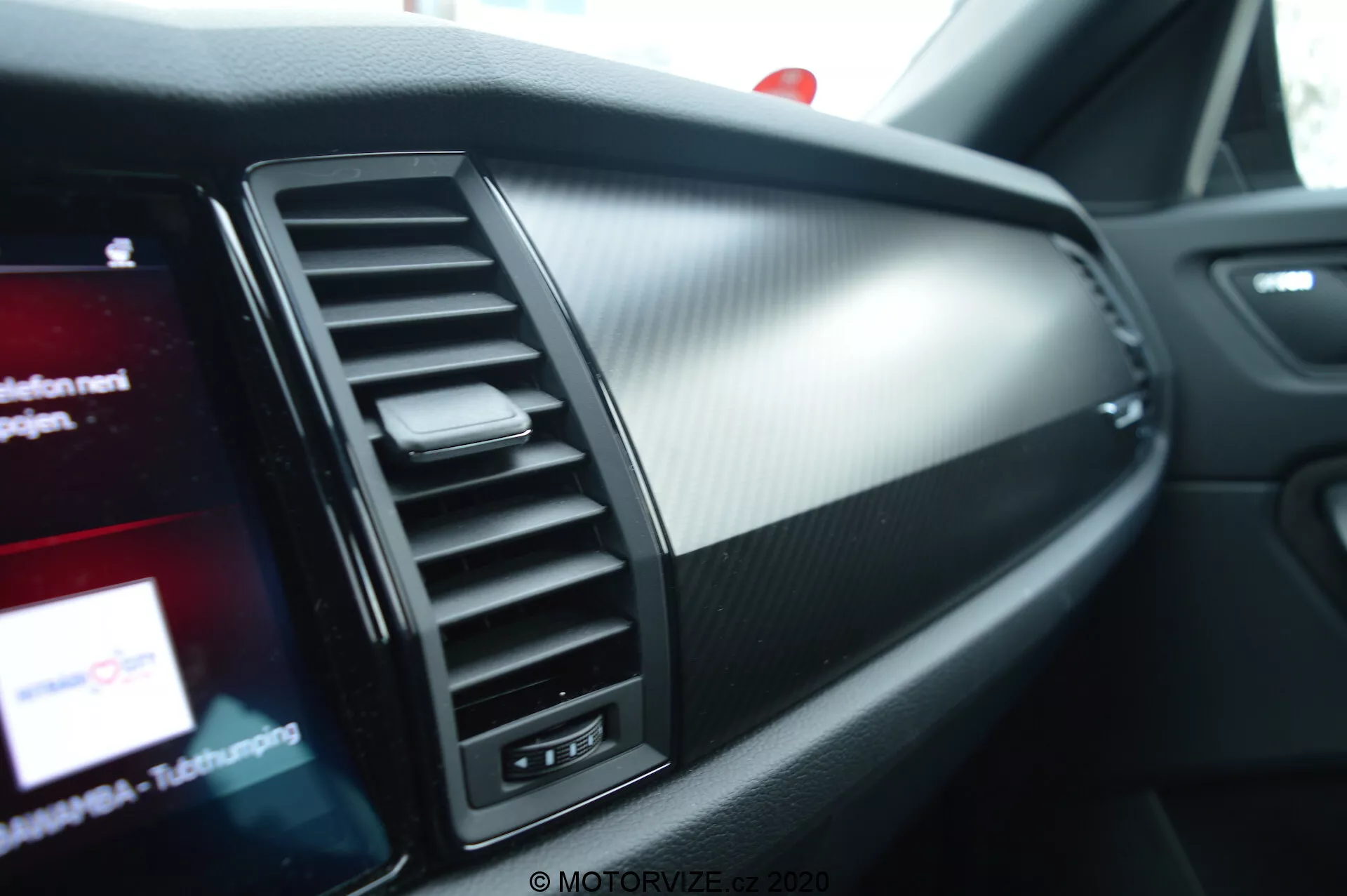 Detailní pohled na interiér Škoda Kodiaq 2017, se zaměřením na spolujezdcovu stranu palubní desky. Obsahuje větrací otvor s vertikálně nastavitelnými lamelami, dekorativní lištu a částečně viditelnou infotainment obrazovku s barevnou grafikou. Materiály palubní desky kombinují kartáčovaný kov a texturovaný plast připomínající karbon, což zdůrazňuje moderní, sportovní design.
