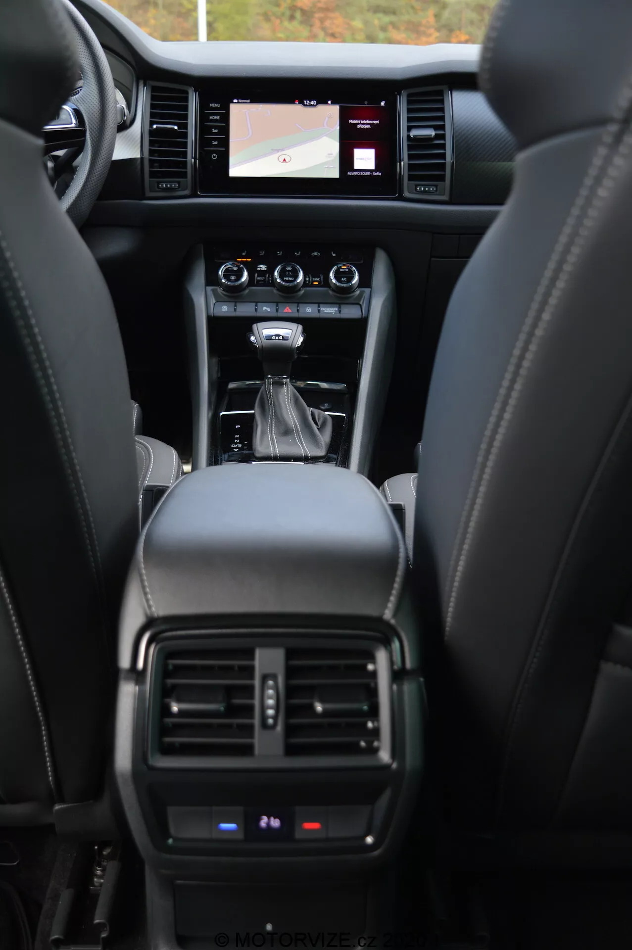 Interiér vozu Skoda Kodiaq 2017, zobrazující přístrojovou desku z pohledu zadních sedadel. Na obrázku jsou kožená sedadla, centrální konzole s automatickou řadicí pákou, ovládací tlačítka, dotykový informační systém zobrazující navigační mapu, ventilační otvory klimatizace a částečný výhled na přístrojový panel zobrazující důležité provozní údaje.