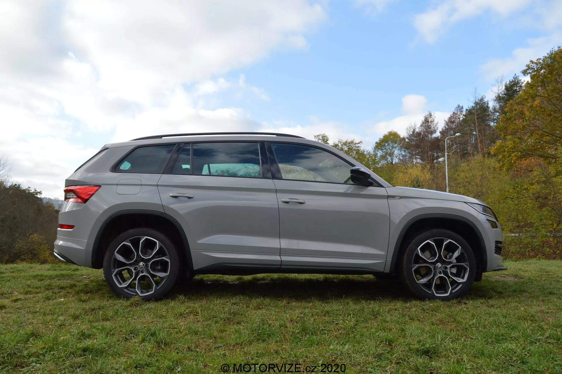 Celkový boční pohled na stříbrný SUV Skoda Kodiaq 2017 zaparkovaný na trávě s pozadím stromů a zatažené oblohy. Design vozidla se vyznačuje výraznými liniemi, velkými slitinovými koly a charakteristickými vícepaprskovými ráfky. Zrcátka, která mají integrované ukazatele směru, jsou namontována těsně pod linií oken. Okna jsou lehce tónovaná a střešní linie se jemně svažuje k zadní části, což naznačuje sportovní, kupé-like profil. SUV je také vybaveno střešním nosičem.
