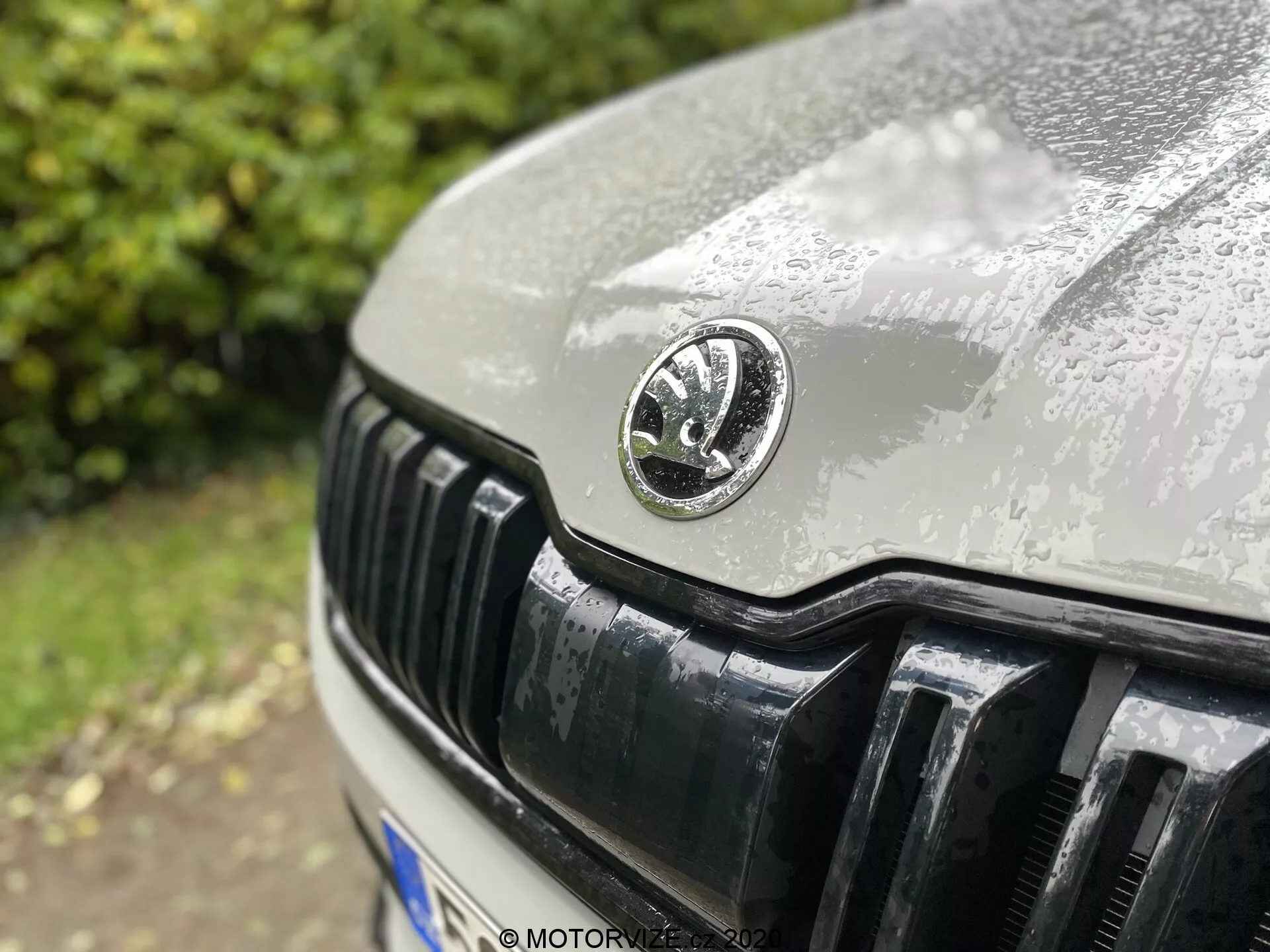 Detailní záběr přední části Škody Kodiaq 2017 zaměřený na znak Škoda, zobrazující lesklou černou mřížku s vertikálními lamelami, kapotu pokrytou kapkami deště a část pravého světla, s rozmazaným pozadím.