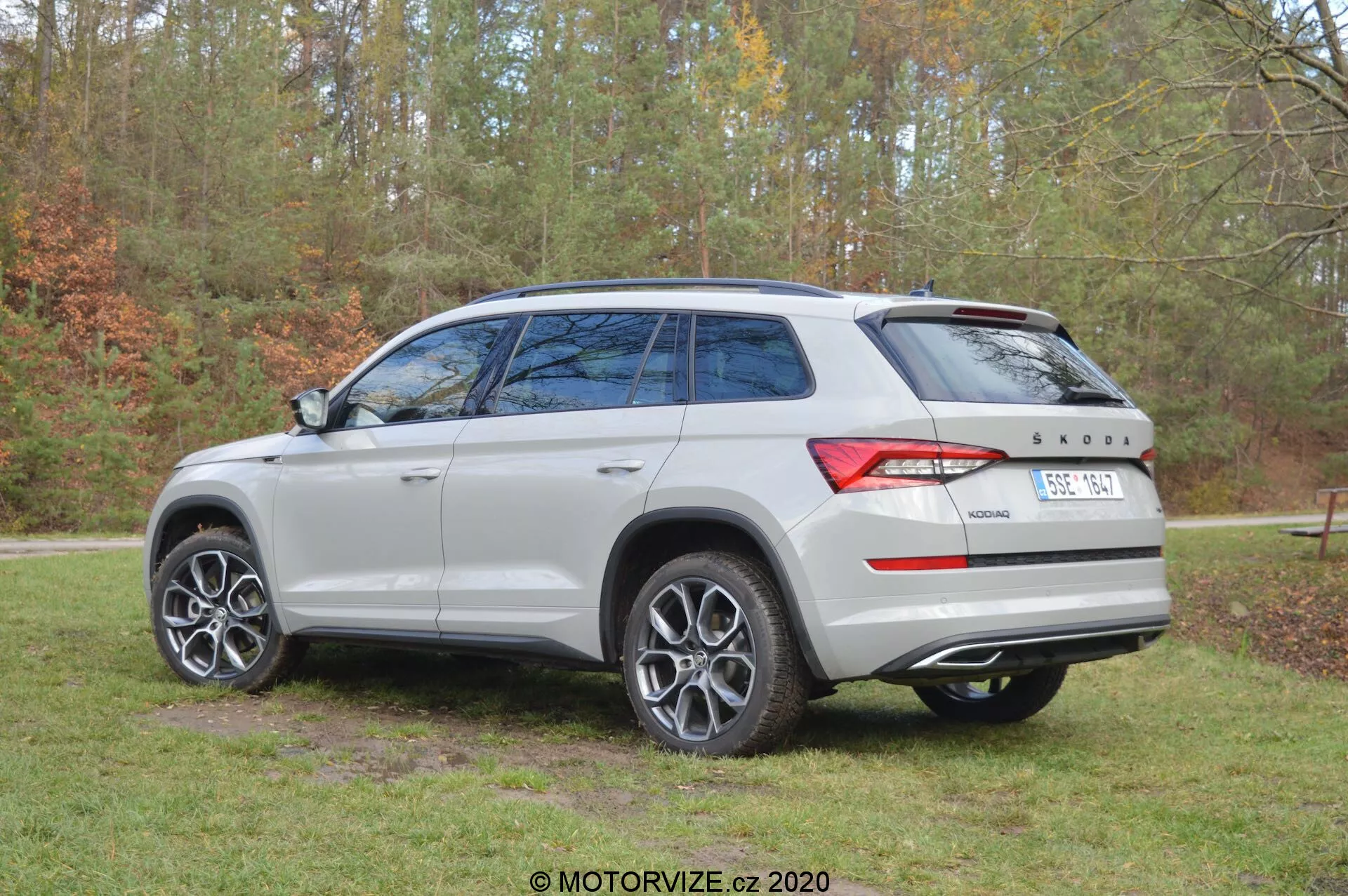 Obrázek Škody Kodiaq 2017 pořízený z tři čtvrtě zadního úhlu, zobrazující stříbrnou/světle šedou barvu vozidla, vícepaprsková kola z lehkých slitin, tónovaná zadní okna, panoramatické střešní okno a charakteristický design zadních světel. Auto je zaparkováno venku na pozadí stromů a trávy, což naznačuje přirozené prostředí. Na zadních dveřích je vidět značka Škoda a znak pohonu '4x4'.