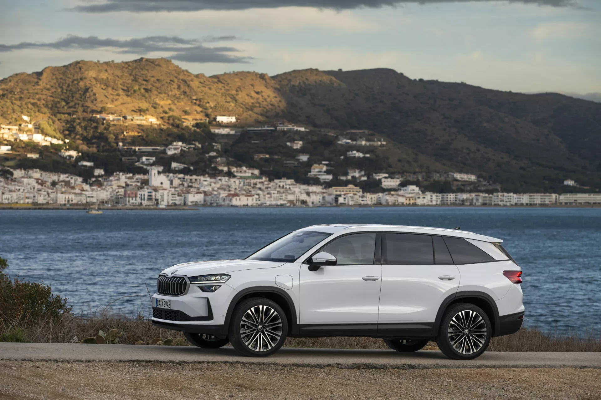 Bílá prémiová SUV Škoda Kodiaq 2024 zaparkovaná na rovné silnici s pobřežní scenérií v pozadí, předvádějící elegantní boční profil vozidla s chromovými detaily a velkými slitinovými ráfky