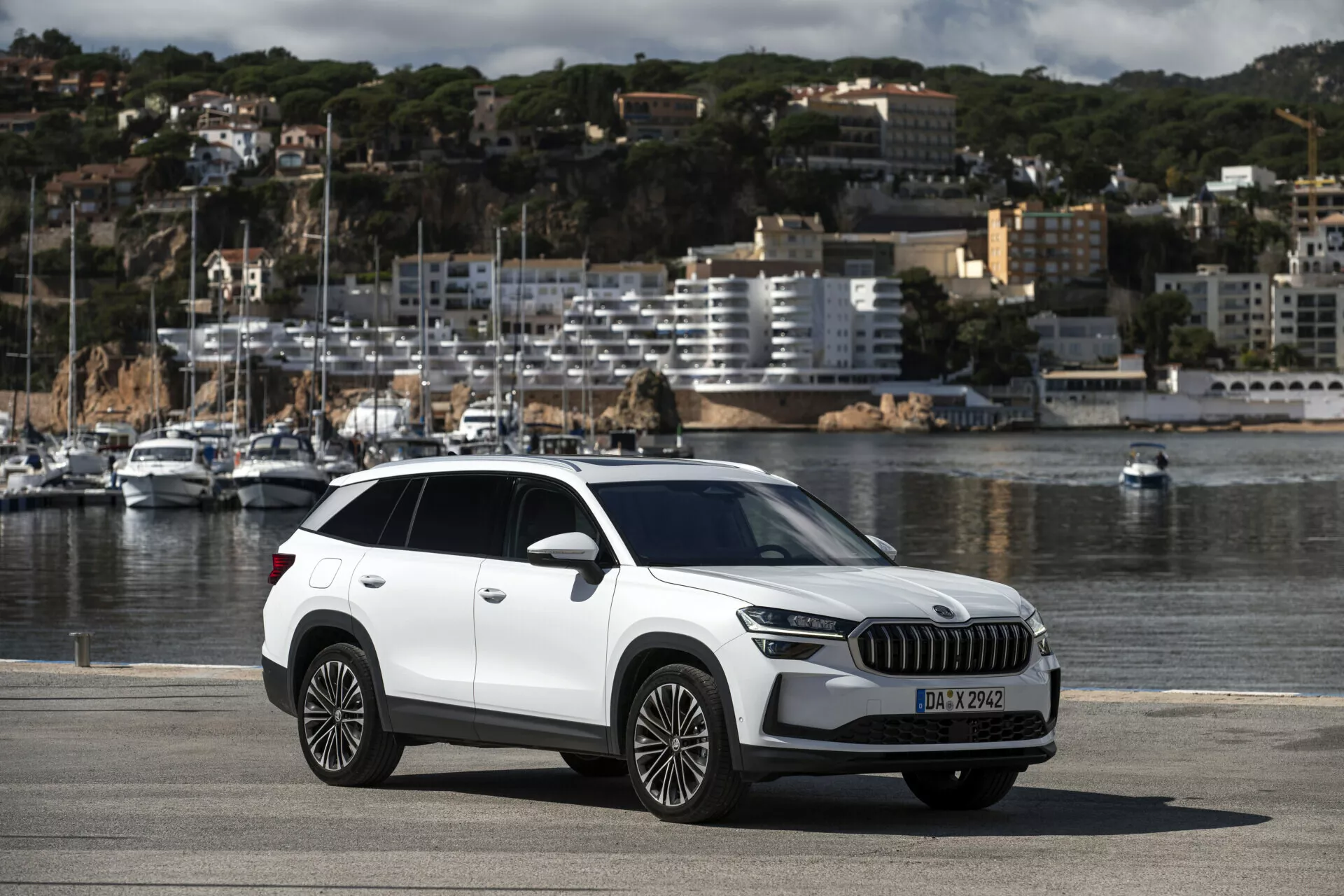Crossover SUV Skoda Kodiaq 2024 zaparkovaný na nábřeží u moře, zobrazující celý vůz včetně moderních prvků jako jsou LED světlomety, velká mřížka, slitinová kola s více paprsky, s malebným pozadím lodí a městské krajiny