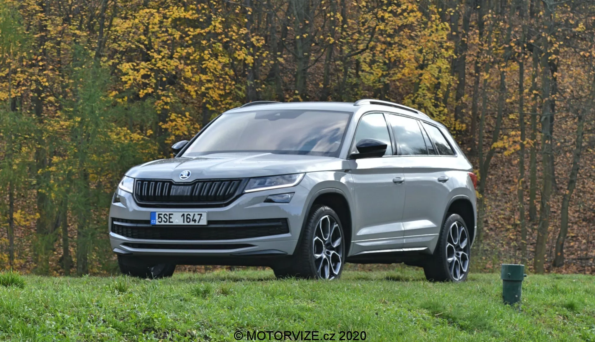 Obrázek zobrazuje SUV Škoda Kodiaq 2017 zaparkované na travnaté ploše s podzimními stromy v pozadí. Vozidlo je foceno z tříčtvrtečního předního pohledu a má horizontální lamelovou mřížku s logem Škoda uprostřed. Elegantní LED světlomety se táhnou do boků a pod nimi se nacházejí mlhovky. Kapota má jemné záhyby a spodní část předního nárazníku zdobí stříbrný lem. Profil vozu zahrnuje vícepaprsková litá kola, zrcátka s integrovanými směrovkami, střešní ližiny a tvarované linie karoserie. Snímače nebo kamery na spodní části nárazníku naznačují pokročilé asistenční systémy řidiče (ADAS). Venkovní prostředí zahrnuje silnici a zelený odpadkový koš na pravém okraji záběru.