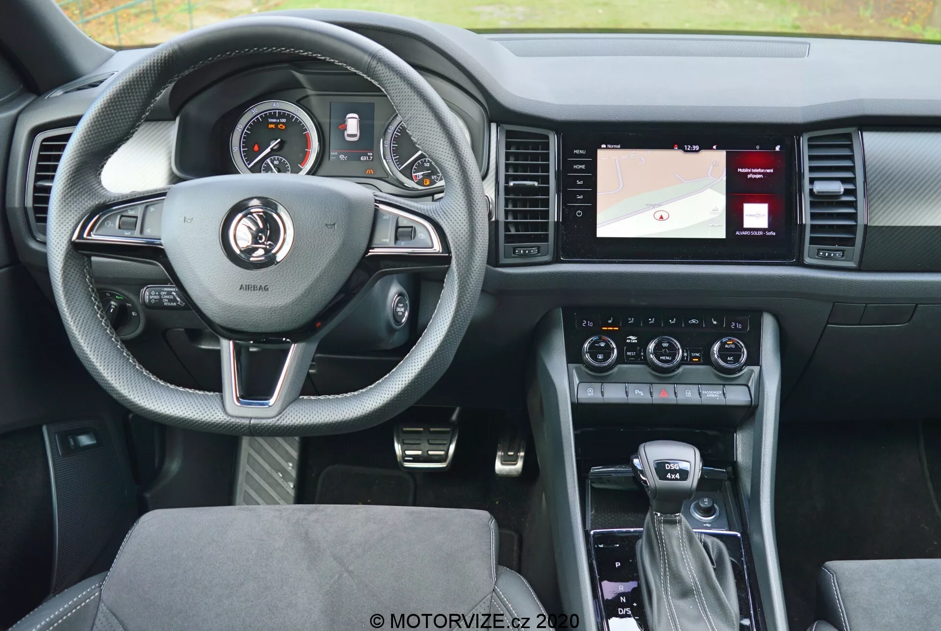 Pohled na interiér vozu Škoda Kodiaq 2017 ze zadního sedadla, zvýrazňující kožený tříramenný volant se stříbrnými akcenty a ovládacími tlačítky pro tempomat, audio a hands-free komunikaci. Přístrojový panel za volantem obsahuje jak analogové ciferníky, tak digitální displej. Středová konzole vpravo zahrnuje obrazovku s navigací, ovládání klimatizace a nastavení audio systému. Řadicí páka naznačuje automatickou převodovku s dalšími ovládacími prvky pro infotainment systém a jízdní režimy. Moderní design směřuje k pohodlí a vyváženosti technologií s čalouněnými sedadly a dobře osvětleným, uživatelsky přívětivým rozvržením.