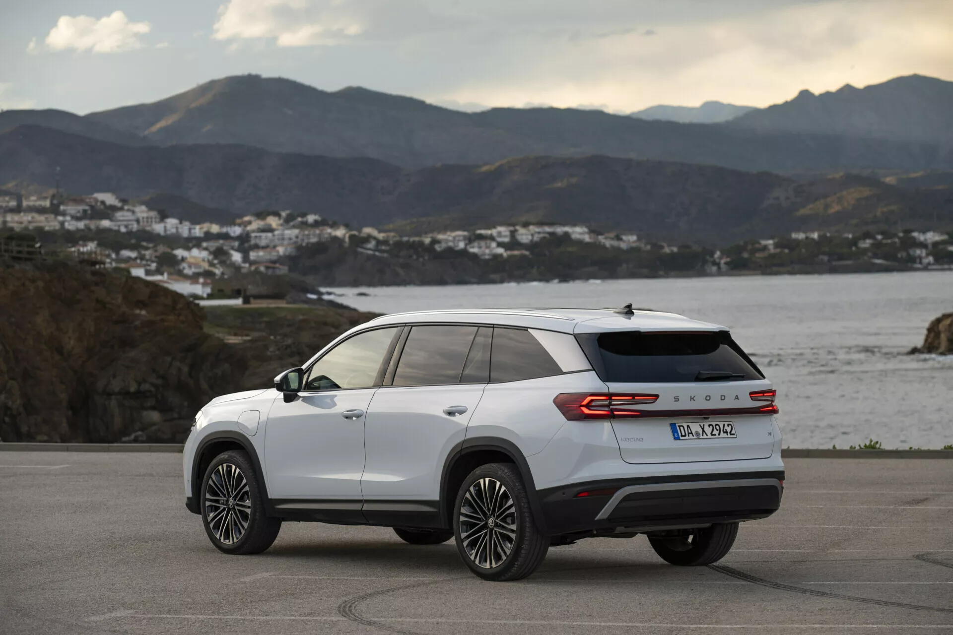 Zadní tříčtvrteční pohled na bílý Skoda Kodiaq 2024, prezentující boční a zadní designové linie, slitinová kola s více paprsky, LED zadní světla a označení modelu, zaparkovaný venku na pobřežní silnici ve zlaté hodině