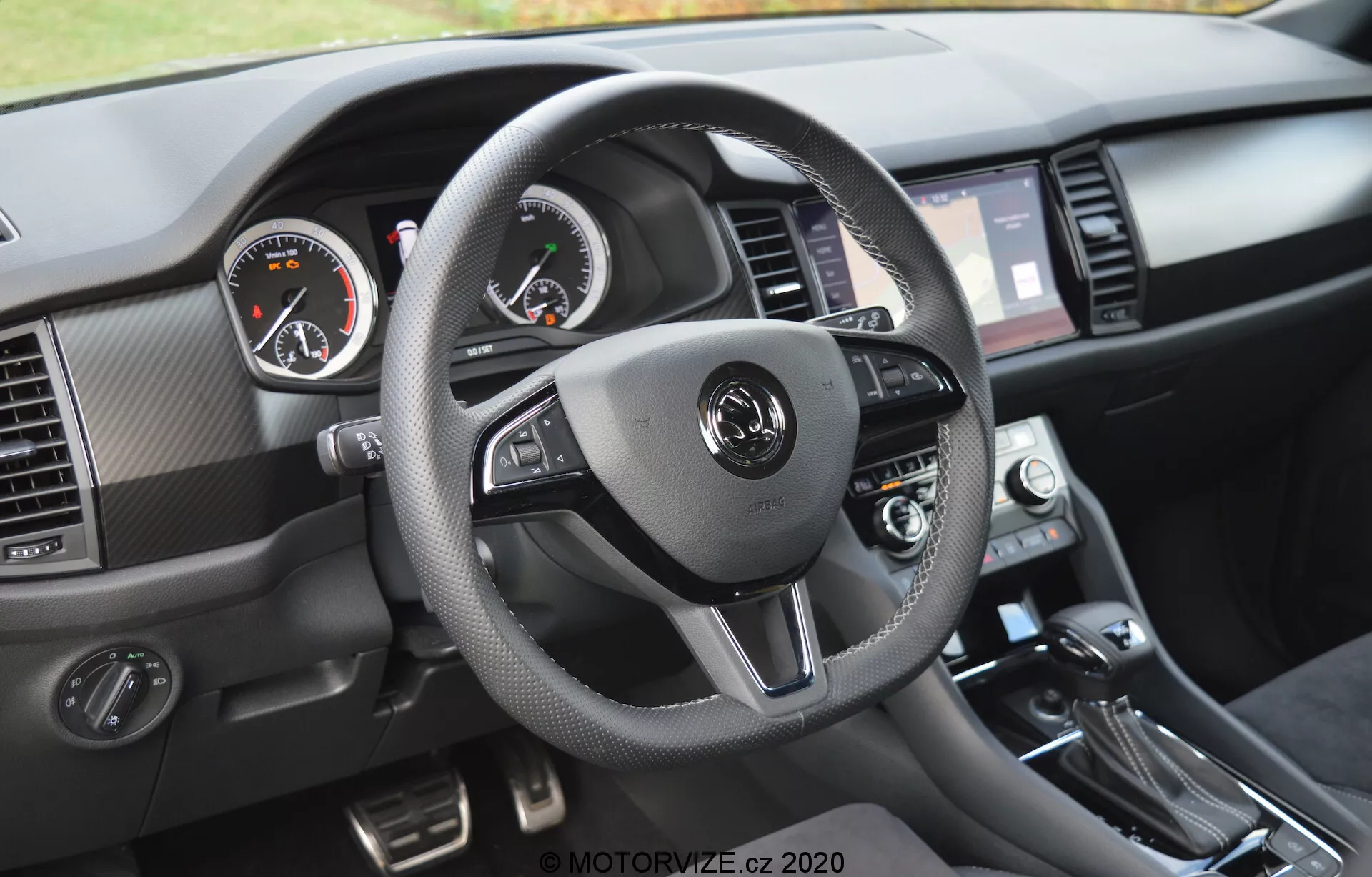 Interiér modelu Škoda Kodiaq 2017 z pohledu řidiče na palubní desku. Tříramenný volant obalený materiálem podobným kůži s kontrastním šitím má integrované ovládací prvky pro audio, hands-free komunikaci a tempomat. Analogové přístroje, s tachometrem vlevo a otáčkoměrem vpravo, doplňuje digitální displej mezi nimi. Viditelné jsou také infotainment dotyková obrazovka lemovaná větracími mřížkami, ovladače klimatizace a řadicí páka, spolu s texturovanými materiály a kartáčovanými kovovými akcenty.
