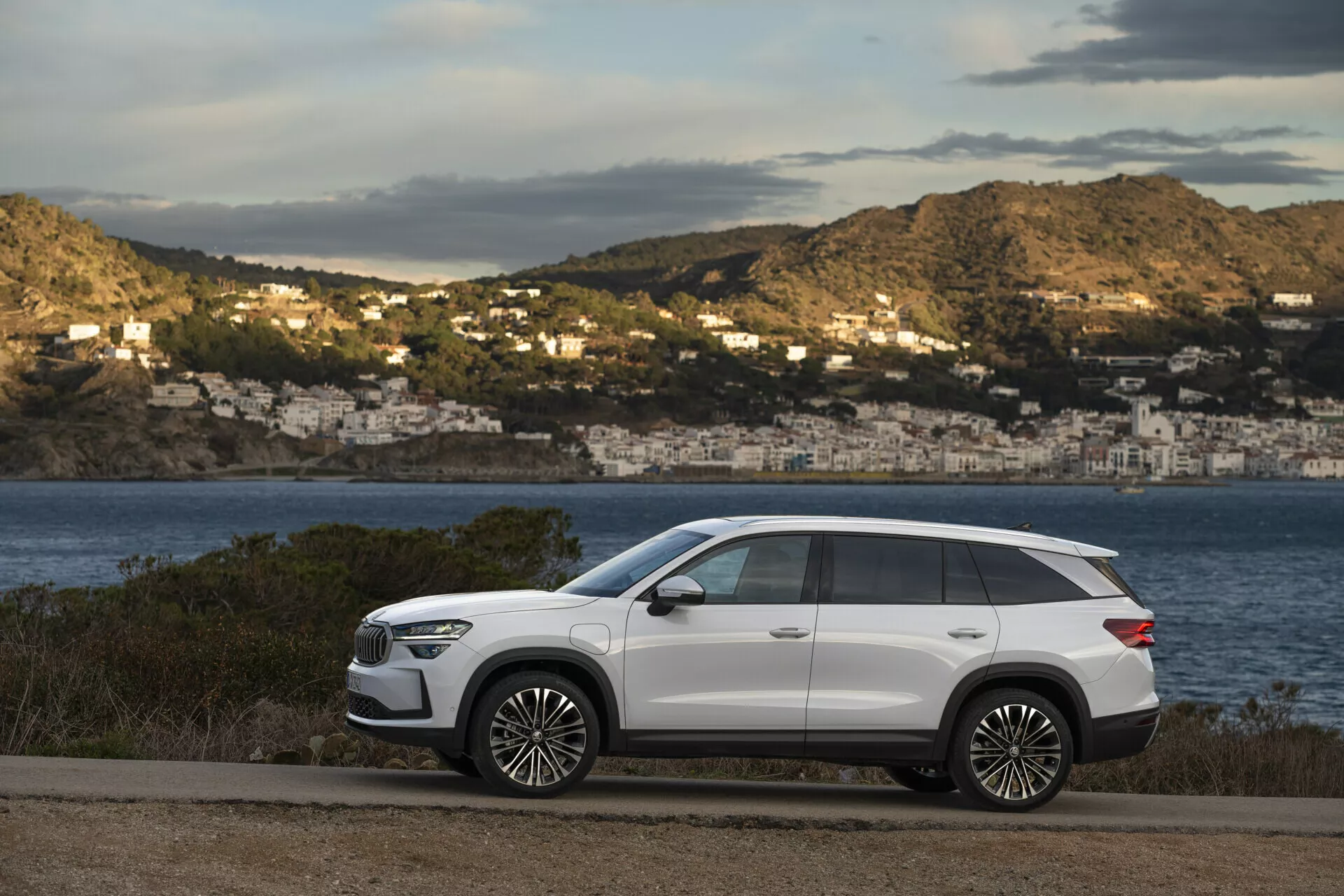 Moderní SUV Skoda Kodiaq 2024 zaparkované na pobřežní silnici s výhledem na malebný záliv a domy na svazích v pozadí, s prvky jako jsou elegantní design, dlouhý rozvor, vysoká světlá výška, ostré linie karoserie, zapuštěné kličky dveří, tónovaná zadní okna, střešní nosiče, velká slitinová kola, LED světlomety, zrcátka se směrovými signály a panoramatické střešní okno, vše v kontrastu s bílou barvou exteriéru.