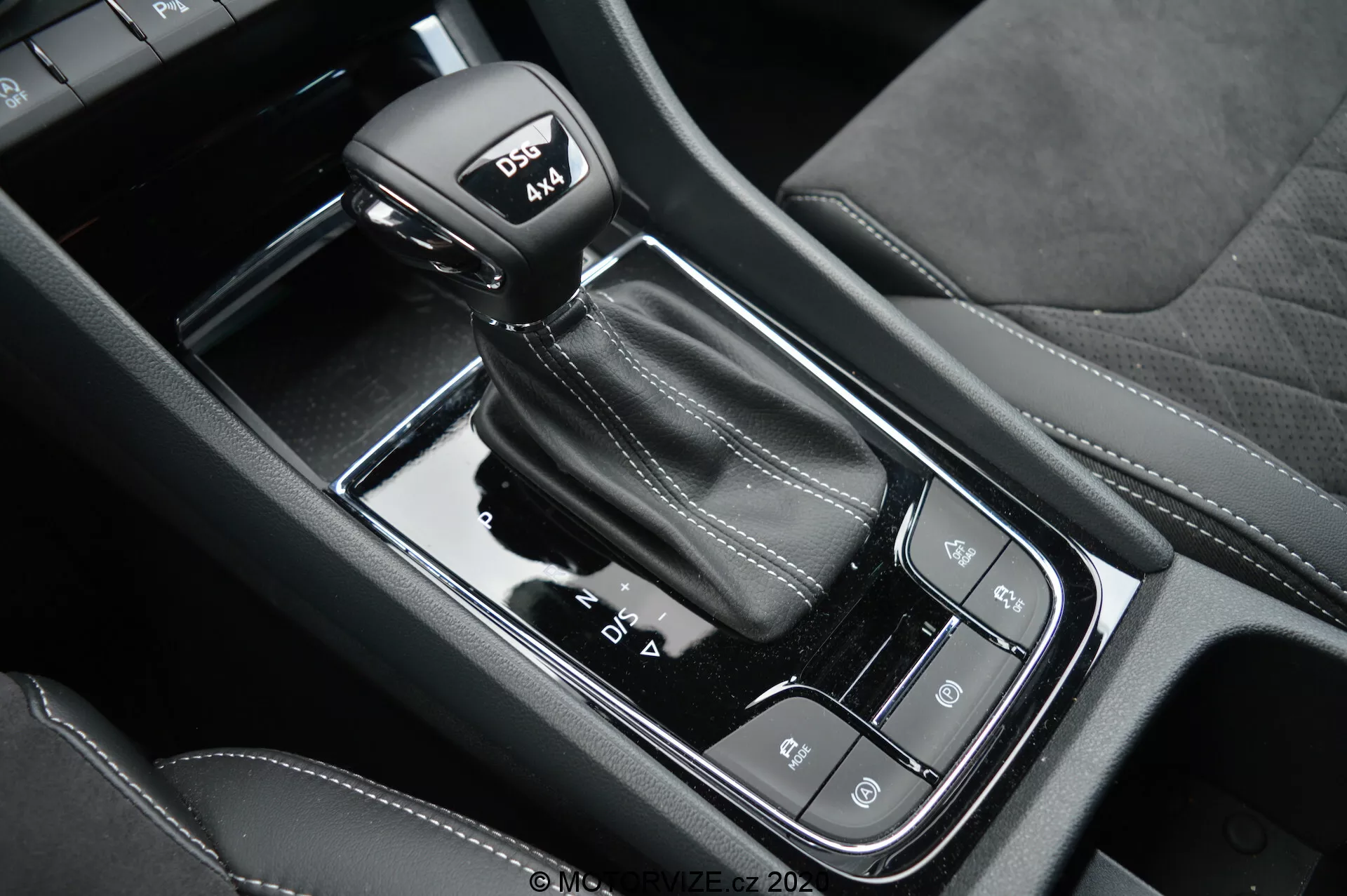 Detailní pohled na automatickou řadicí páku a lesklou černou středovou konzoli vozu Skoda Kodiaq 2017, s funkcemi jízdních režimů, elektronické parkovací brzdy a tlačítkem auto-hold; zdůraznění koženého prošívání na knoflíku umožňujícím sportovní jízdu.