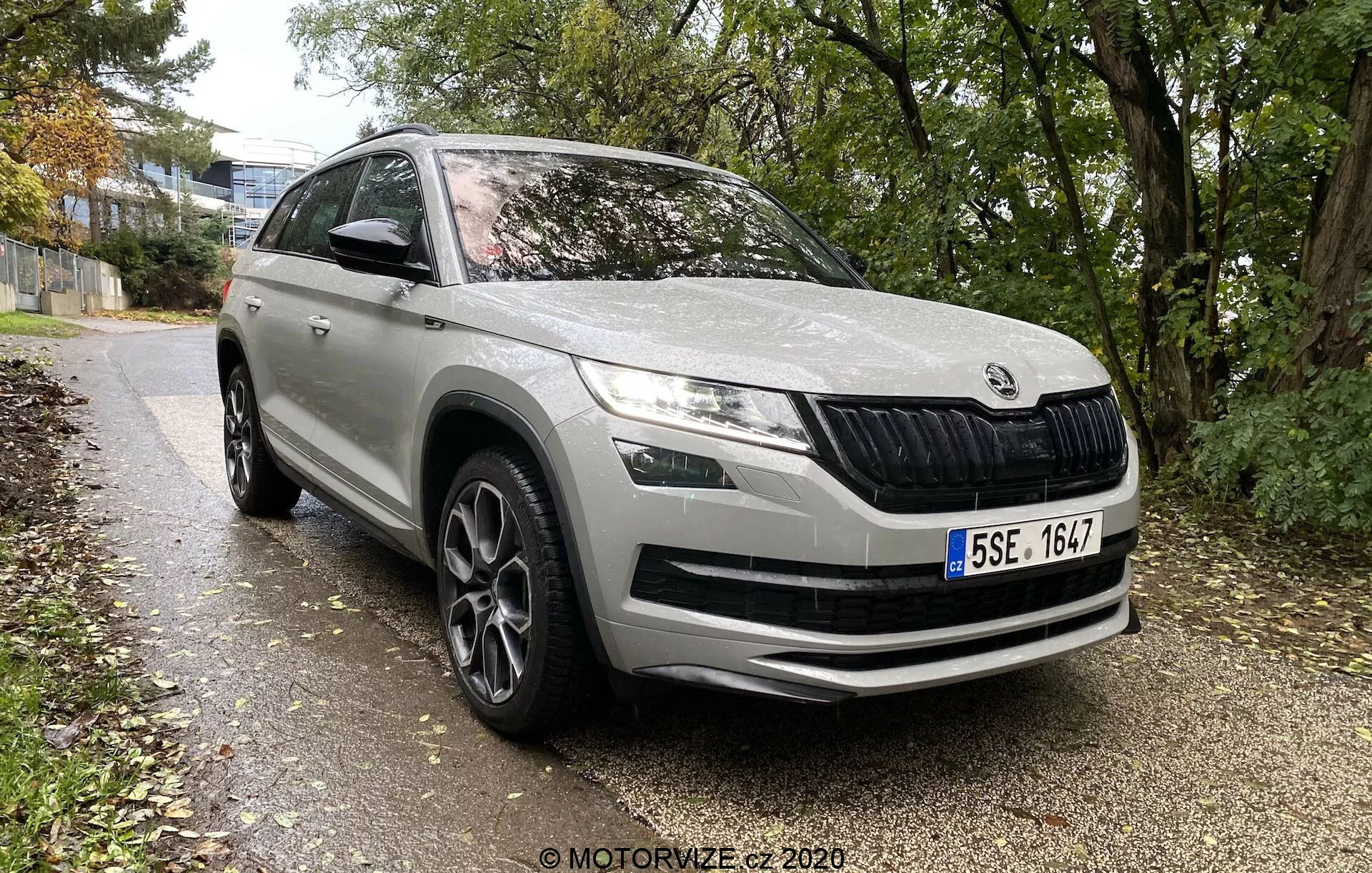 Škoda Kodiaq 2017 moderní SUV od evropské automobilky, zepředu zezadu ve třech čtvrtinách ukazuje přední a boční profil až do poloviny zadních dveří, zaparkované podél cesty lemované trávou a stromy v městském nebo předměstském prostředí. Vlastnosti zahrnují charakteristickou přední mřížku s vertikálními lamelami, elegantní LED světlomety, mlhovky v nárazníku, velké vzduchové otvory na předním nárazníku, kontury kapoty pro aerodynamiku, výrazné podběhy kol s vícepaprskovými koly z lehkých slitin, boční zrcátka s integrovanými směrovkami, ochranné lišty na spodní straně a celkově čistý, dobře udržovaný lak.