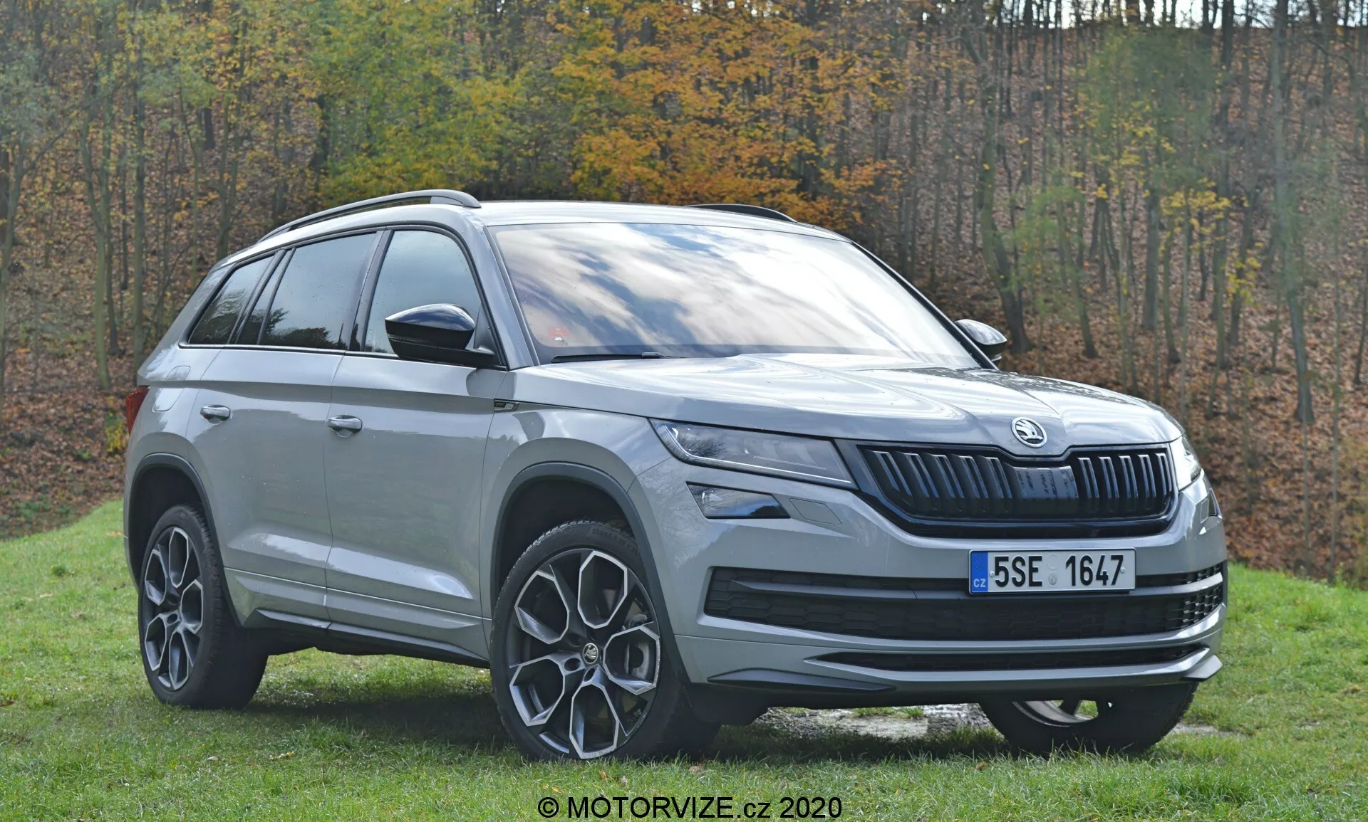 Tento obrázek zobrazuje SUV Škoda Kodiaq z roku 2017 zachycené z úhlu zboku zepředu, zaměřující se na levou stranu vozidla. Snímek zvýrazňuje přední mřížku s charakteristickým svisle šachovaným designem Škoda, konfiguraci světlometů pravděpodobně s LED technologií a středové logo značky. Boční profil zahrnuje přední dveře u řidiče, zadní dveře u spolujezdce, střešní ližiny a části zadní čtvrtiny. Také vynikají nápadná hliníková kola s paprskovým designem. Venkovní prostředí se zatravněným pozadím a podzimním listím zvyšuje estetiku moderního SUV.
