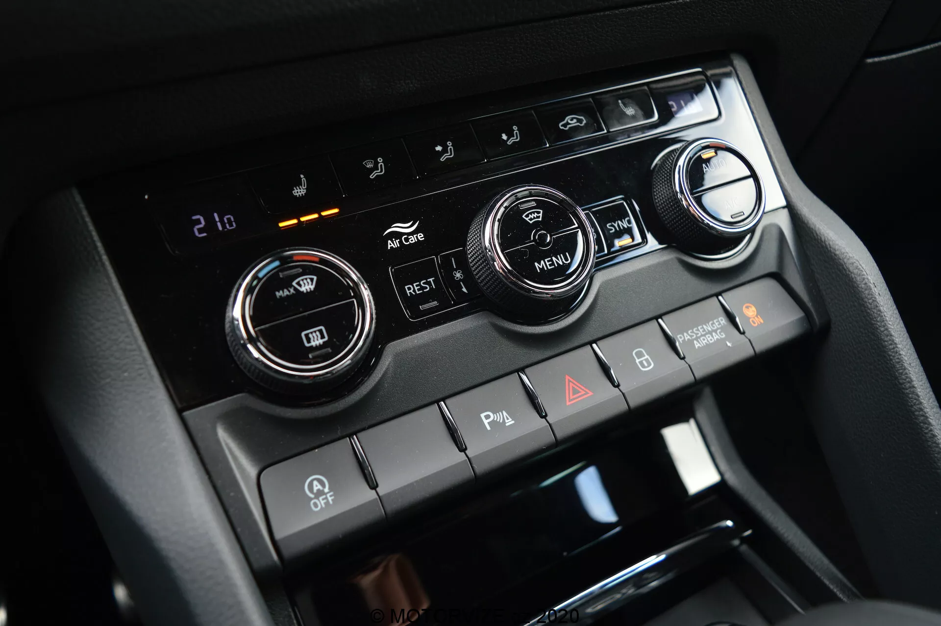 Detailní pohled na středovou konzolu Škoda Kodiaq 2017, která zvýrazňuje systém regulace klimatu se dvěma velkými otočnými knoflíky pro nastavení teploty a digitálním displejem nastaveným na 21 stupňů Celsia. Obrázek také obsahuje tlačítka pro klimatizaci, recirkulaci vzduchu, odmlžování oken a tlačítko 'MENU'. Níže jsou přepínače pro parkovací senzory, deaktivaci automatického start/stop a výstražná světla.