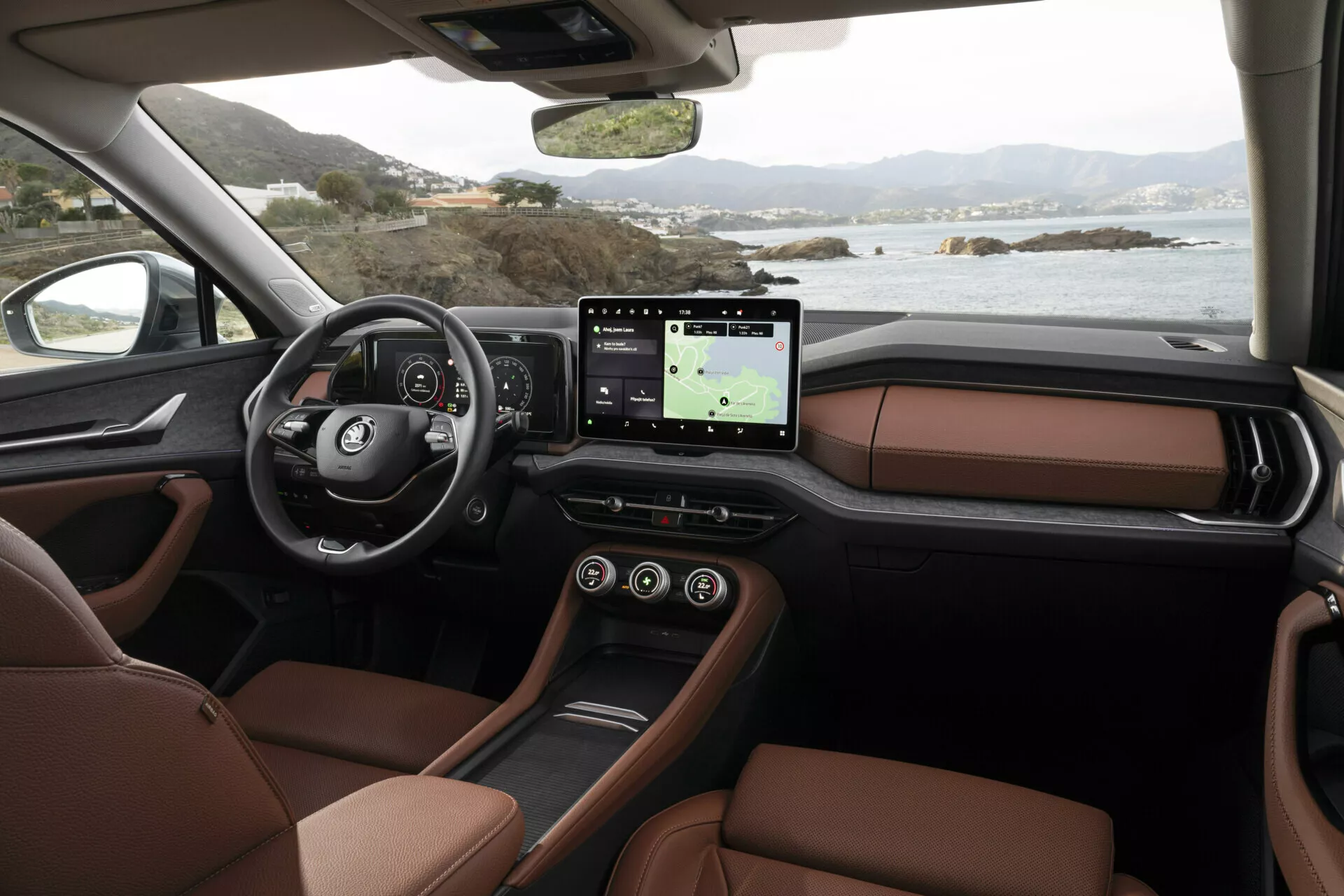 Interiér Škoda Kodiaq 2024 z pohledu spolujezdce zobrazující palubní desku, multifunkční volant s logem, digitální přístrojový panel, velký dotykový infotainment systém s GPS, luxusní hnědá kožená sedadla a elegantní design středové konzole, s výhledem na klidnou pobřežní krajinu skrze okna