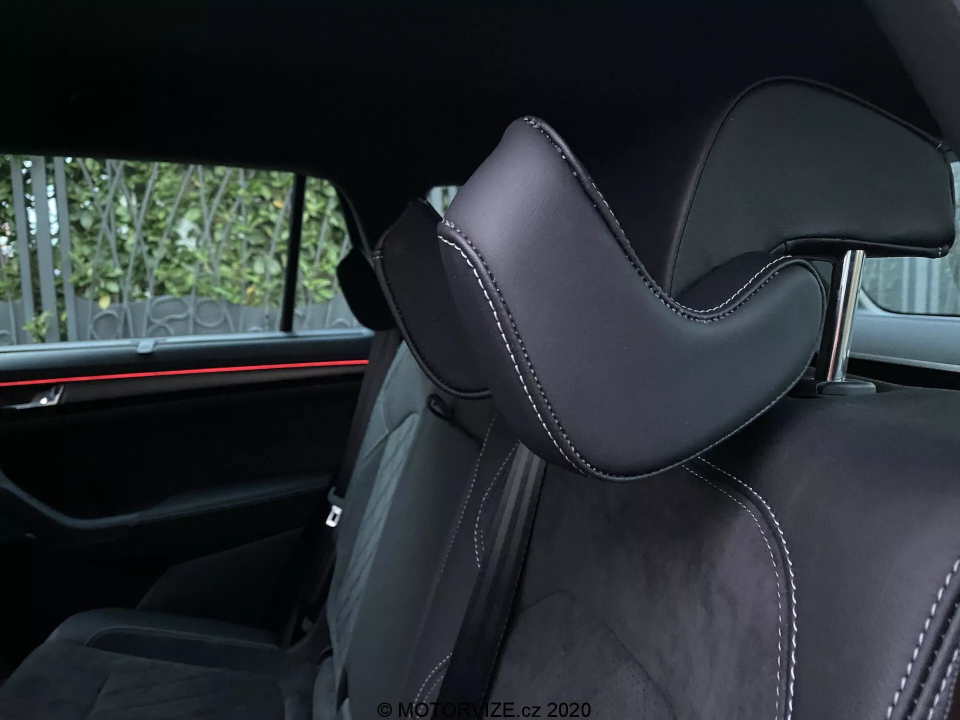 Interiér vozu Skoda Kodiaq 2017 zaměřený na autosedačku. Úhel záběru zdůrazňuje část opěrky hlavy, čalouněnou černým materiálem, pravděpodobně kůží nebo vysoce kvalitním syntetickým materiálem, s výrazným tvarováním pro pohodlí. Precizní prošívání kontrastní nití přidává luxusní detaily. Na pozadí je vidět horizontální červené ambientní osvětlení, které zdůrazňuje moderní prvky automobilového designu. Viditelné zadní pasažérské vybavení zahrnuje bezpečnostní pásy a možná také středovou opěrku, což naznačuje druhou řadu sedadel.