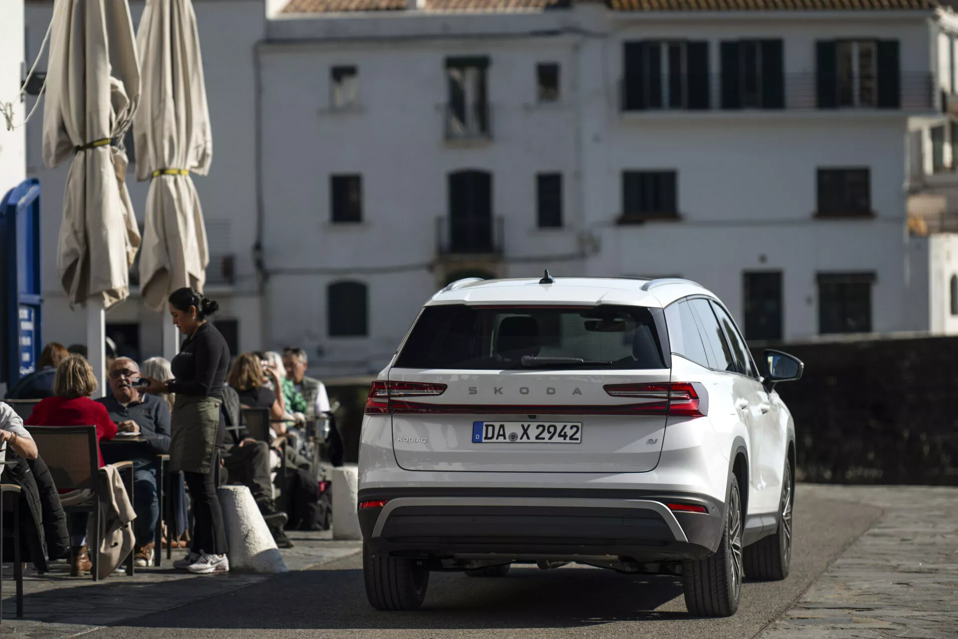 Bílý Škoda Kodiaq 2024 fotografován z mírně zdviženého zadního úhlu, ukazující celou zadní část, zadní světla, víko kufru, zadní okno s stěračem a střechu s anténou a nosiči, zaparkovaný na dlážděné ulici před kavárnou s klasickou evropskou architekturou v pozadí.
