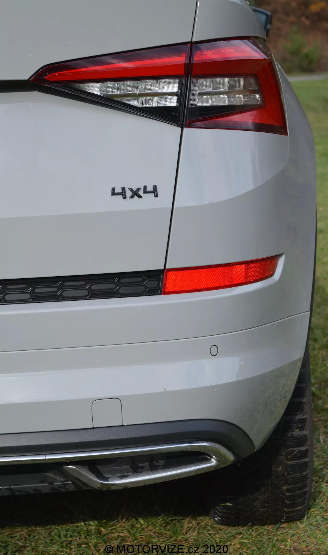 Detailní pohled na zadní roh vozu Skoda Kodiaq 2017, zaměřený na zadní osvětlovací soustavu, nápisy a design nárazníku. Úhel záběru zdůrazňuje pravou stranu zadní části vozu, která ukazuje horizontálně uspořádané, ostré, hranaté zadní světlo obklopující bok. Pod zadním světlem je vidět zadní čtvrtpanel a nárazník v barvě stříbrné nebo světle šedé, který odráží tlumené prostředí. Nárazník obsahuje černou plastovou spodní část s voštinovým vzorem a stříbrným nebo chromovým lemem, pravděpodobně souvisejícím s parkovací asistencí nebo stylistikou. Nápis '4X4' nad zadním světlem označuje čtyřkolový pohon. Vysoce kvalitní snímek odhaluje detailní textury povrchu, částečně viditelný běhoun pneumatiky na dně, postavený na trávě. Vodoznak '© MOTORVIZE.cz 2020' naznačuje, že obrázek je určen pro české automobilové publikace.