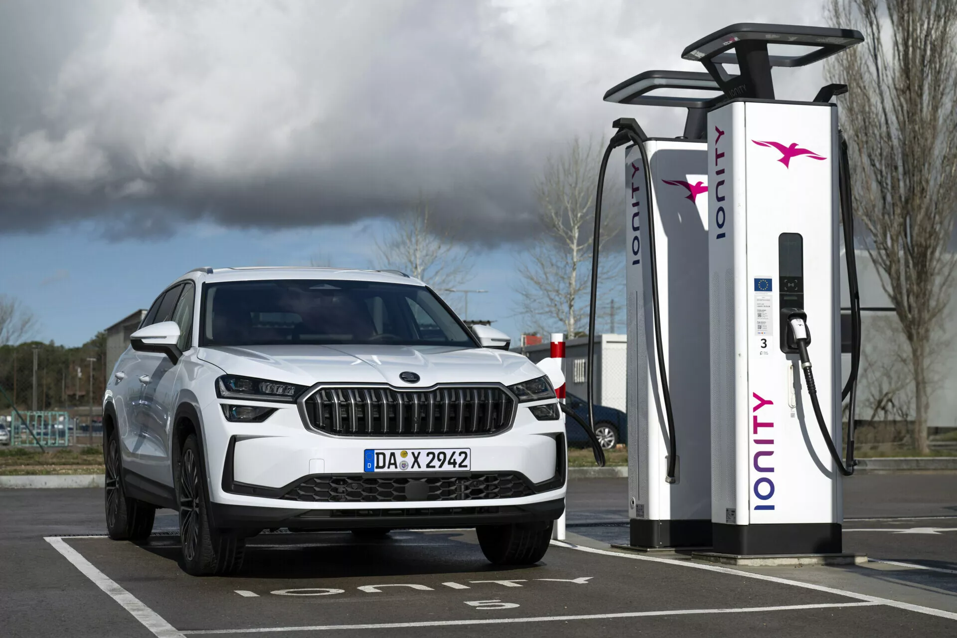 Bílé SUV Skoda Kodiaq 2024 zaparkované u nabíjecí stanice, s pohledem na přední část vozu s LED světlomety a denními světly, zdůrazňující jeho elektrické nebo hybridní možnosti a luxusní design mřížky.