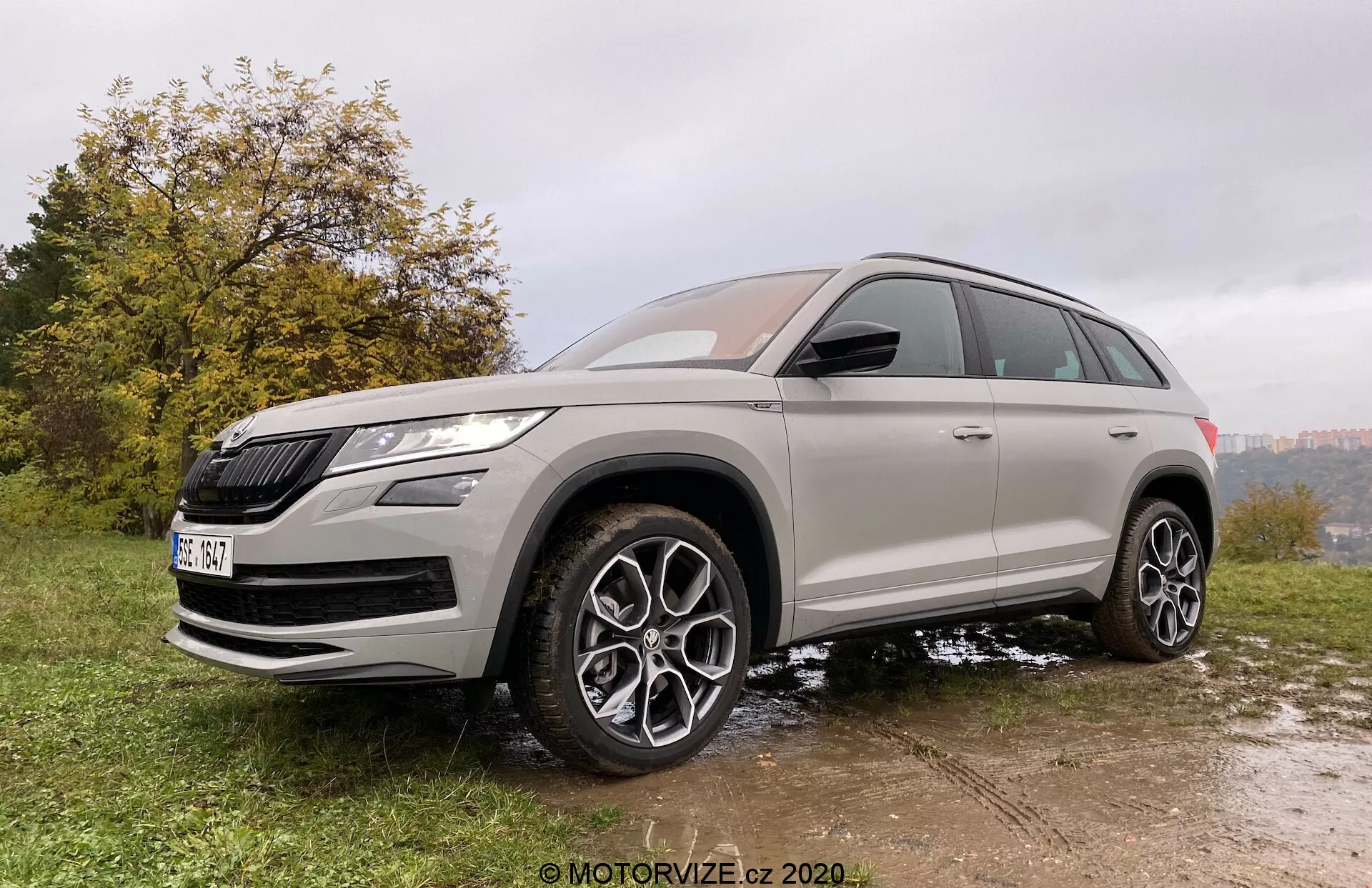 Tříčtvrteční přední pohled na SUV Škoda Kodiaq 2017 pořízený z mírně nízkého úhlu, zdůrazňující výšku a postoj vozidla. Vozidlo je zaparkované na nerovném povrchu z trávy a bláta, což zdůrazňuje jeho off-road schopnosti. Škoda Kodiaq se vyznačuje výraznou maskou chladiče a ostrými LED světlomety. Precizní design zahrnuje velká vícepaprsková litá kola, vysokou světlou výšku a celosezónní pneumatiky. Karoserie je lakována neutrální metalickou barvou s tónovanými okny a chromovými akcenty, které zvýrazňují její prémiový vzhled. Moderní funkce zahrnují zrcátka v barvě karoserie s integrovanými směrovkami a pravděpodobně i systémy varování před mrtvým úhlem.