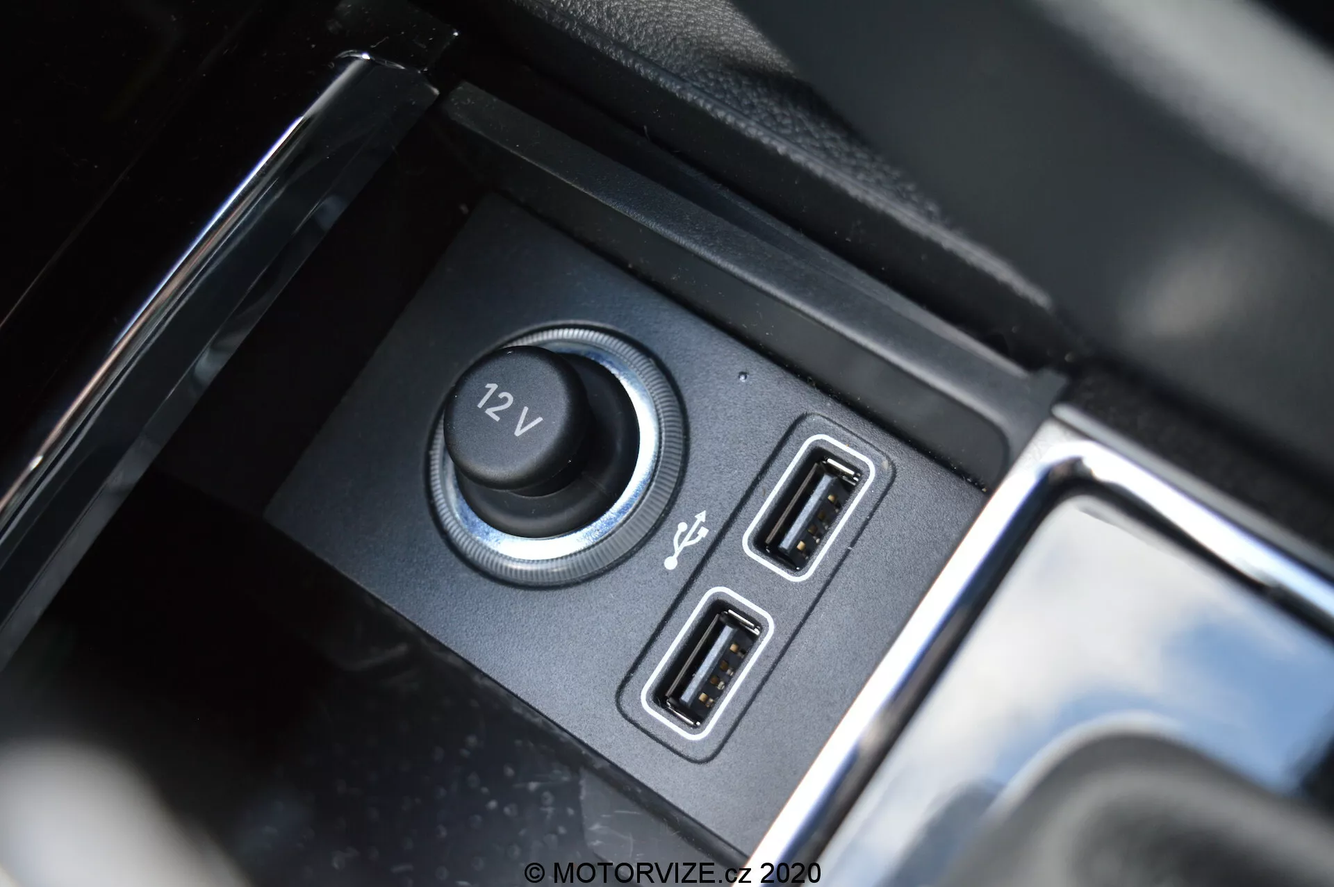 Detailní záběr interiéru Škoda Kodiaq 2017 zaměřený na napájecí a konektivní funkce, včetně 12V zásuvky a dvou USB portů, s matnou černou přihrádkou a lesklými reflexními akcenty.