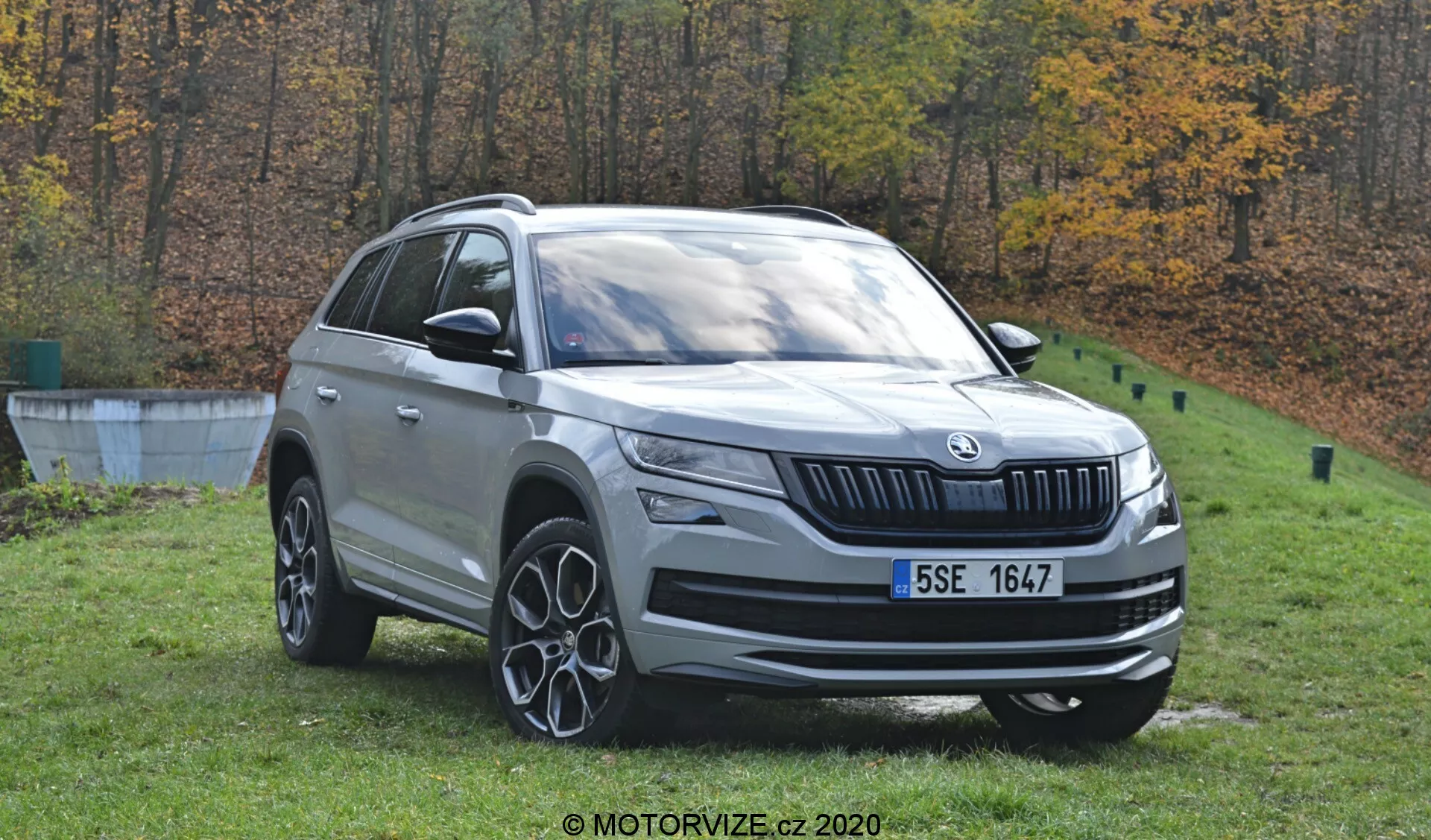 Detailní pohled na Škoda Kodiaq 2017, stříbrné SUV, zasazené venku na podzimním pozadí stromů. Denní snímek zachycuje vozidlo z předního třičtvrtinového úhlu, ukazuje jeho přední a boční profil. Viditelné jsou výrazné designové prvky, včetně přední masky s logem, LED světlometů, mlhovek, vícepaprskových slitinových kol, střešních ližin a zpětných zrcátek s integrovanými směrovkami. Auto je postavené na trávě poblíž betonového okraje, což naznačuje parklike prostředí.