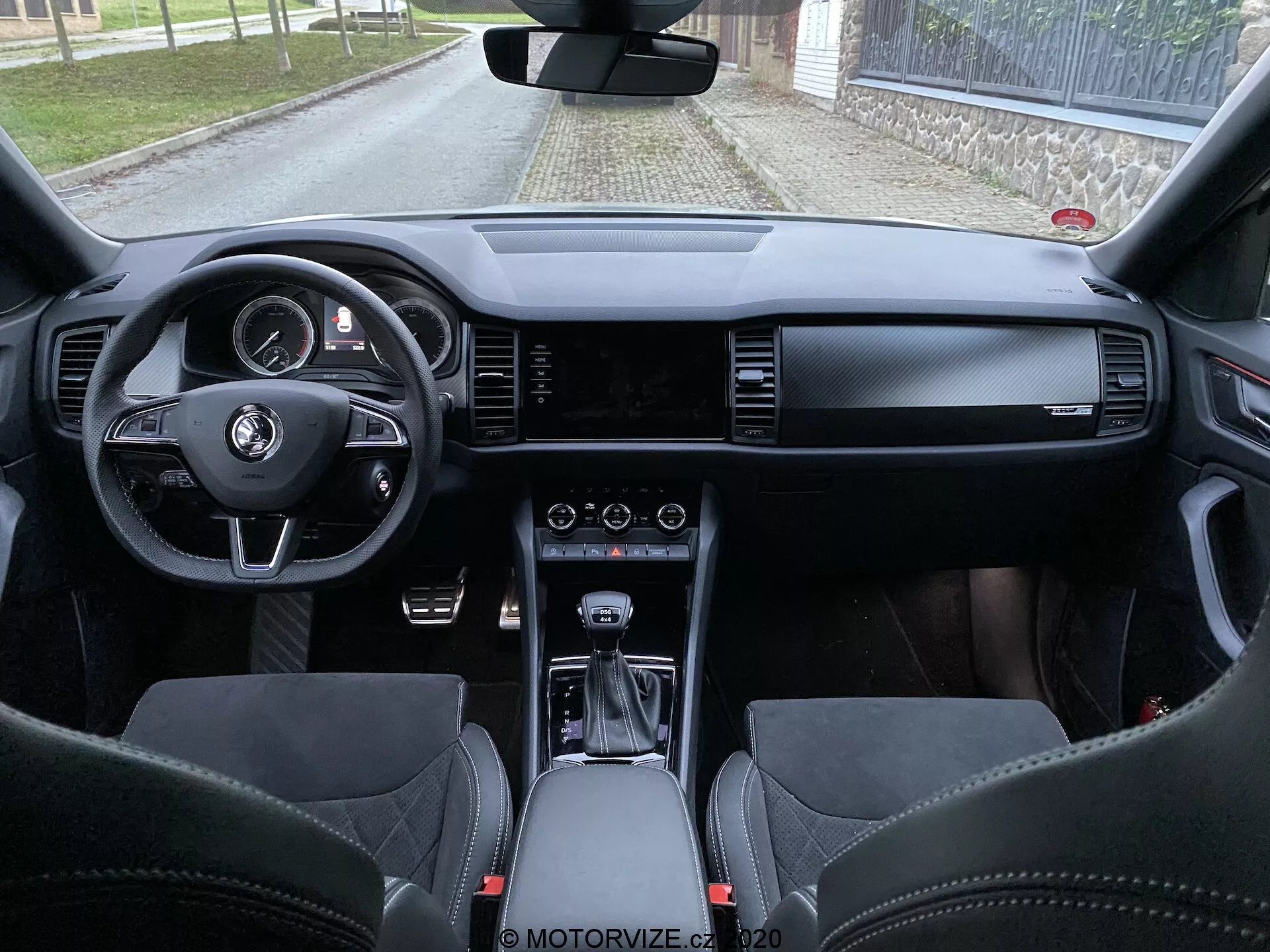 Interiér vozu Škoda Kodiaq 2017 z pohledu zadních sedaček, který ukazuje palubní desku, volant s multifunkčními tlačítky, digitální displej přístrojové desky, široký infotainmentový displej, ovládání HVAC, volič automatické převodovky, prémiové kožené materiály a sportovní sedačky s integrovanými opěrkami hlavy.