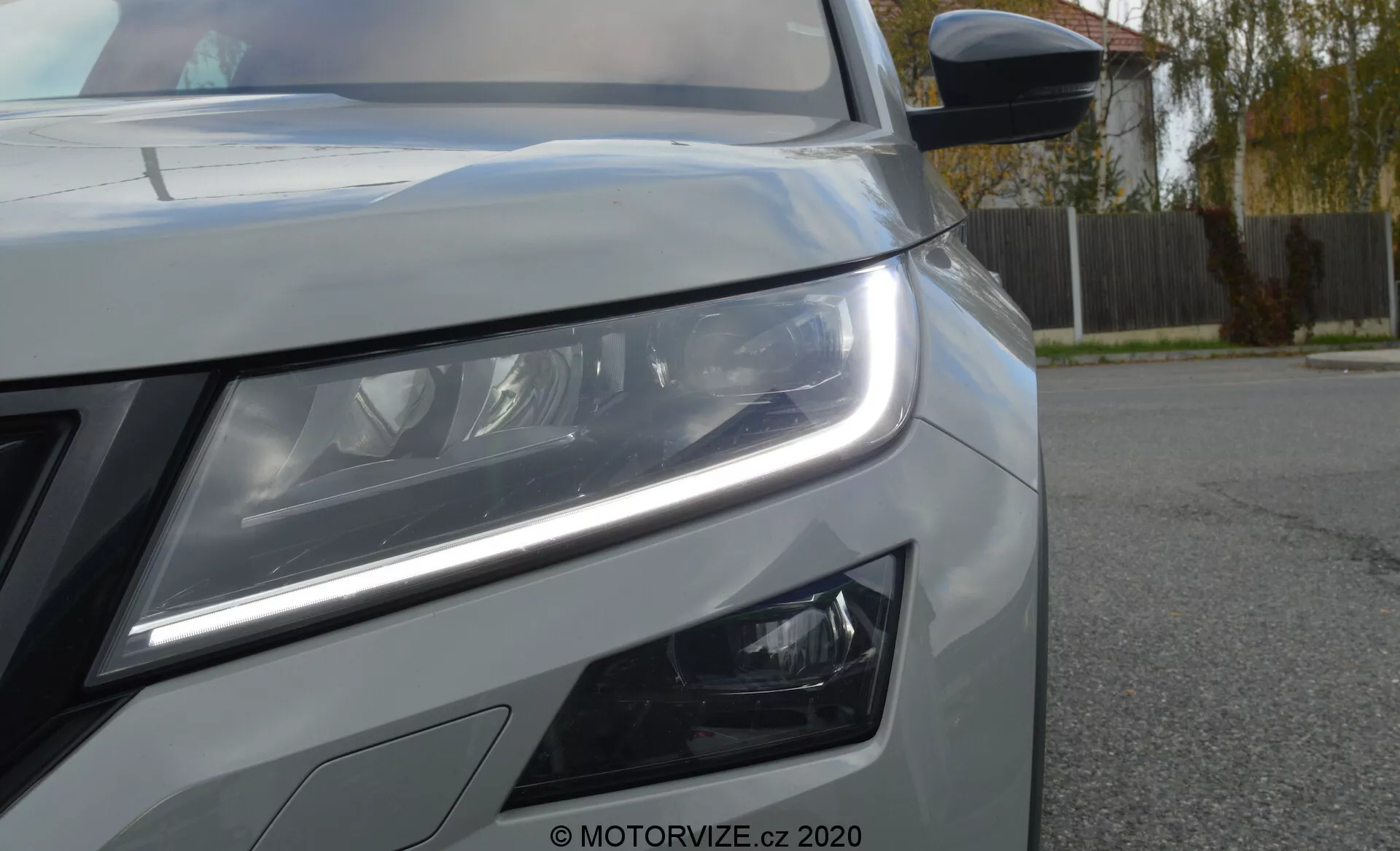 Detailní pohled na přední levý bok vozu Škoda Kodiaq 2017. Nízký úhel zvýrazňuje výrazné LED denní svícení a detailní hlavní světlomety naznačující pokročilou osvětlovací technologii. Pod světlometem jsou viditelné moderně navržené mlhové světlomety. Na obrázku je také část přední mřížky, metalický lak s ostrými liniemi karoserie a boční zrcátko s integrovanou směrovkou. Vodotisk 'MOTORVIZE.cz 2020' označuje zdroj fotografie.
