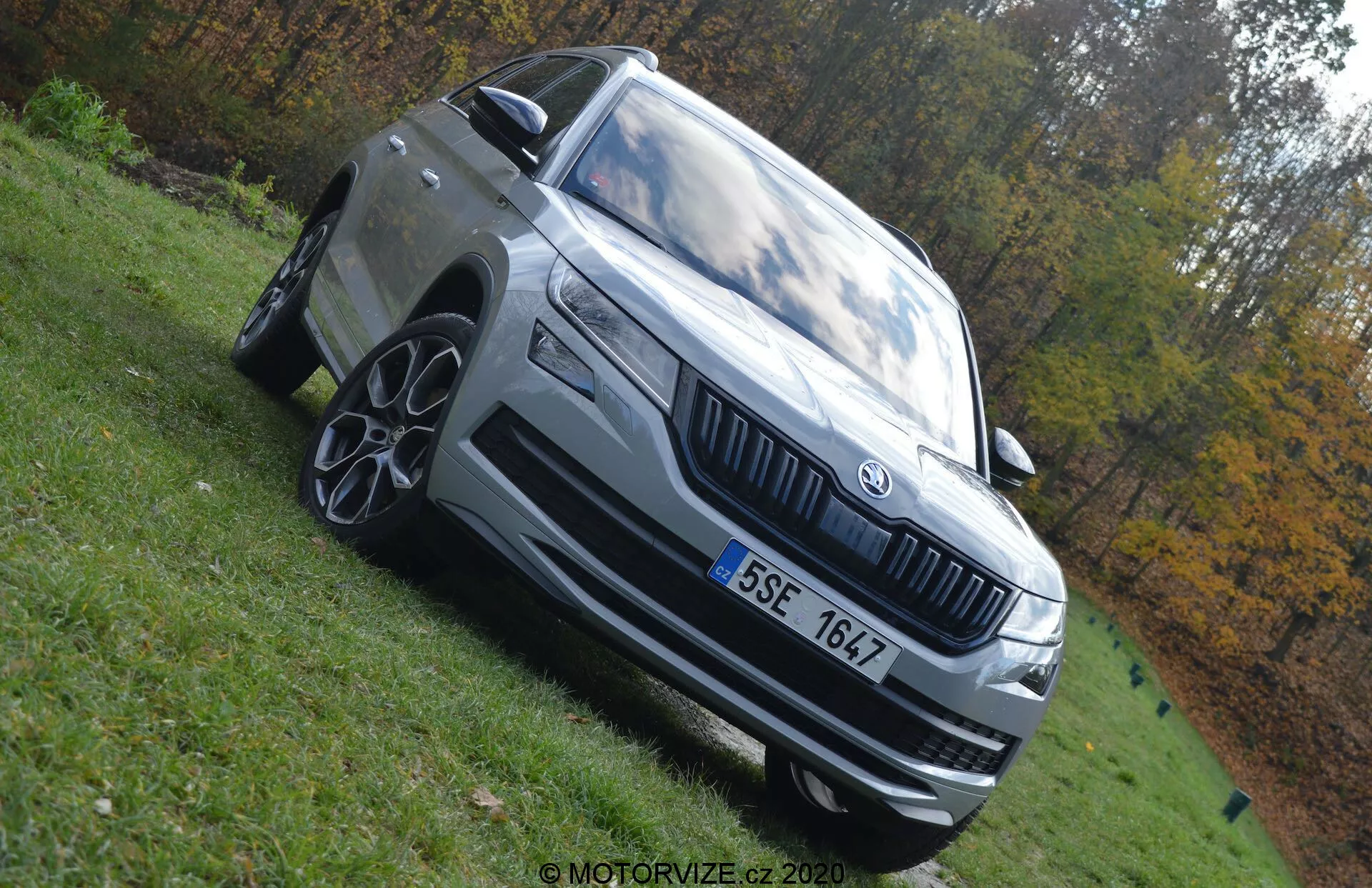 Fotografie vozu Skoda Kodiaq 2017 z nízkého úhlu, která zdůrazňuje jeho dominantní postoj. Auto je zachyceno z levé přední strany s viditelnými prvky, jako jsou LED světlomety, výrazná maska s emblémem Skoda, vícepaprsková litá kola, přední nárazník s nasávacími otvory, boční zrcátko s integrovaným blinkrem a charakteristické linie karoserie. Vozidlo je světle šedé nebo stříbrné a je zaparkováno na travnaté ploše za denního světla. Rozmazané podzimní prostředí podtrhuje přítomnost vozidla.
