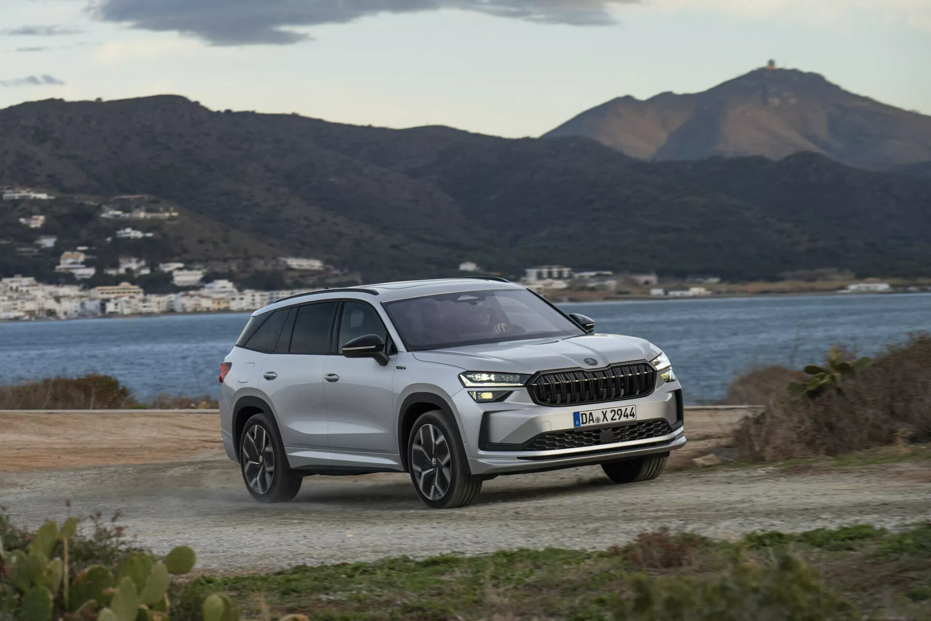SUV Skoda Kodiaq 2024 parkované na nezpevněném povrchu s výhledem na scénické kopce a vodní plochu, zdůrazňující moderní LED světlomety vozu, přední mřížku s potenciální integrací ADAS a současný design kol pod měkkým, zataženým denním světlem.
